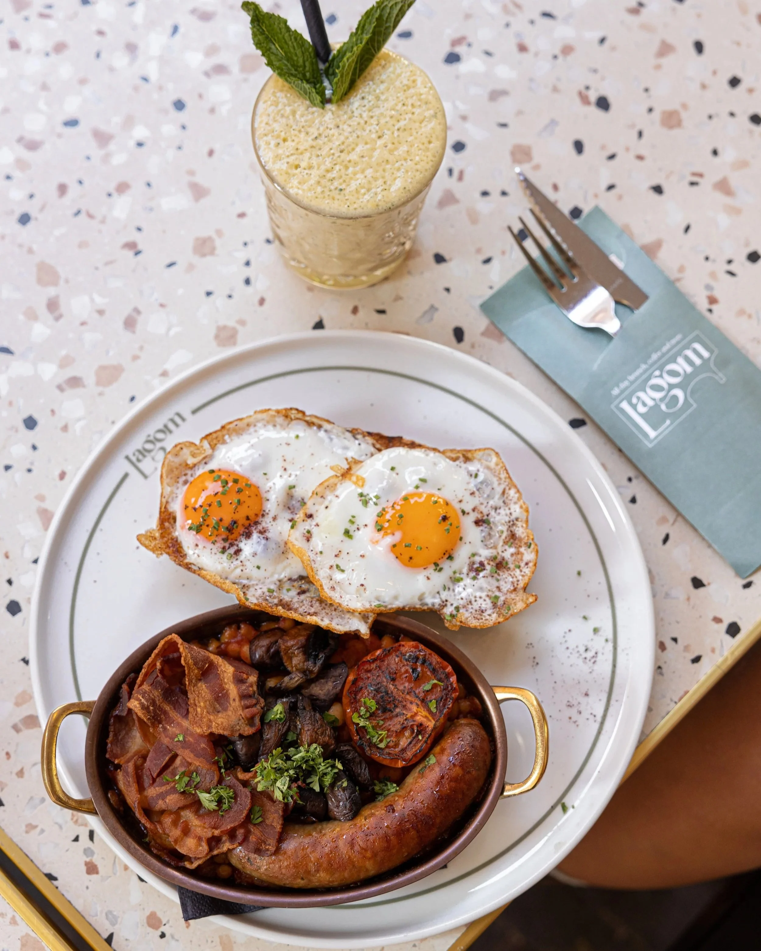 A breakfast plate with sunny side up eggs, a sausage, bacon, grilled mushrooms, and a sausage link in a small, oval-shaped, brown serving dish. The dish is garnished with chopped parsley.