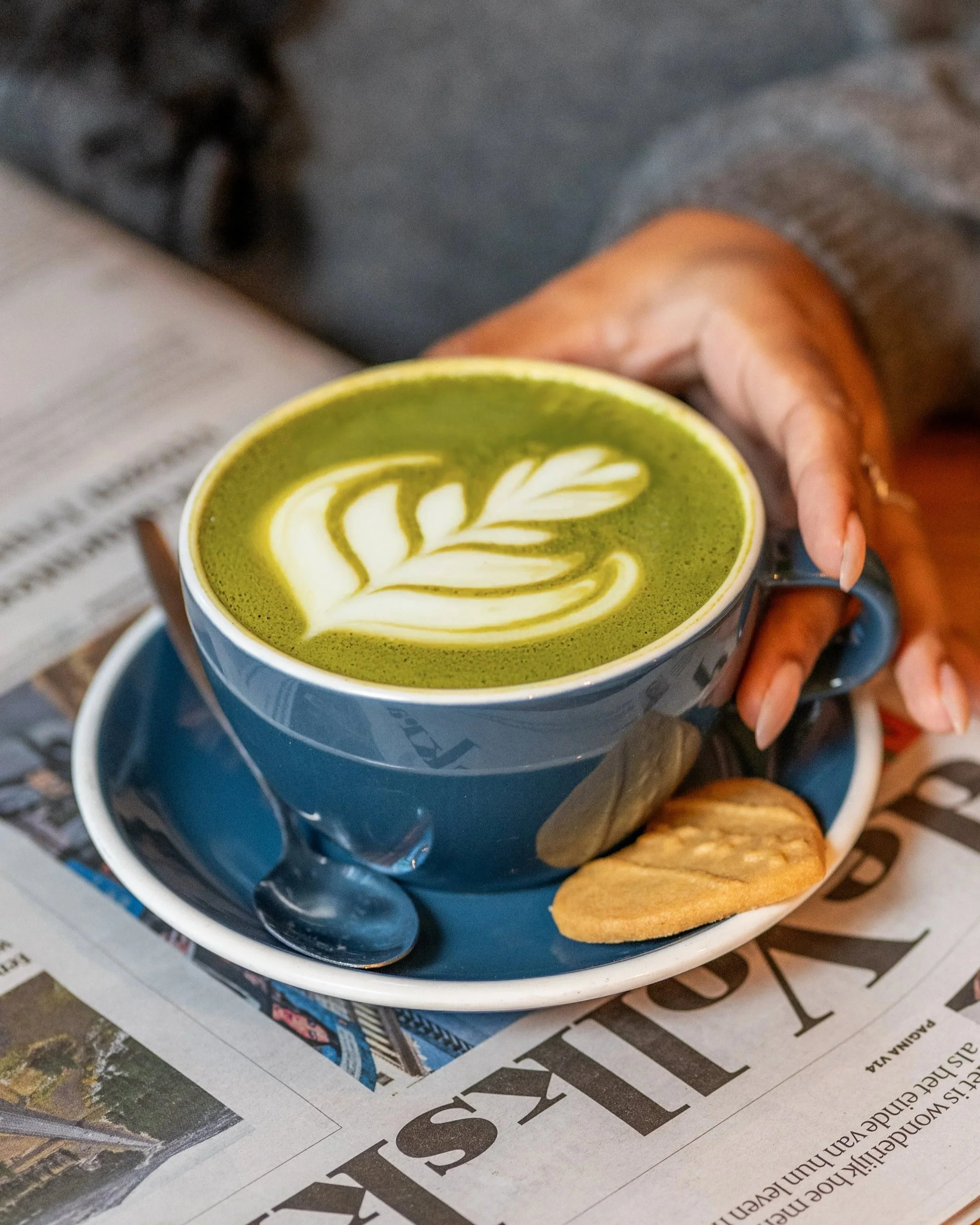 A blue cup of matcha latte with white leaf latte art, resting in a blue saucer, with two cookies beside it, on a newspaper surface. 
Matcha latte with leaf latte art in blue ceramic cup at Lagom Amsterdam all-day brunch café