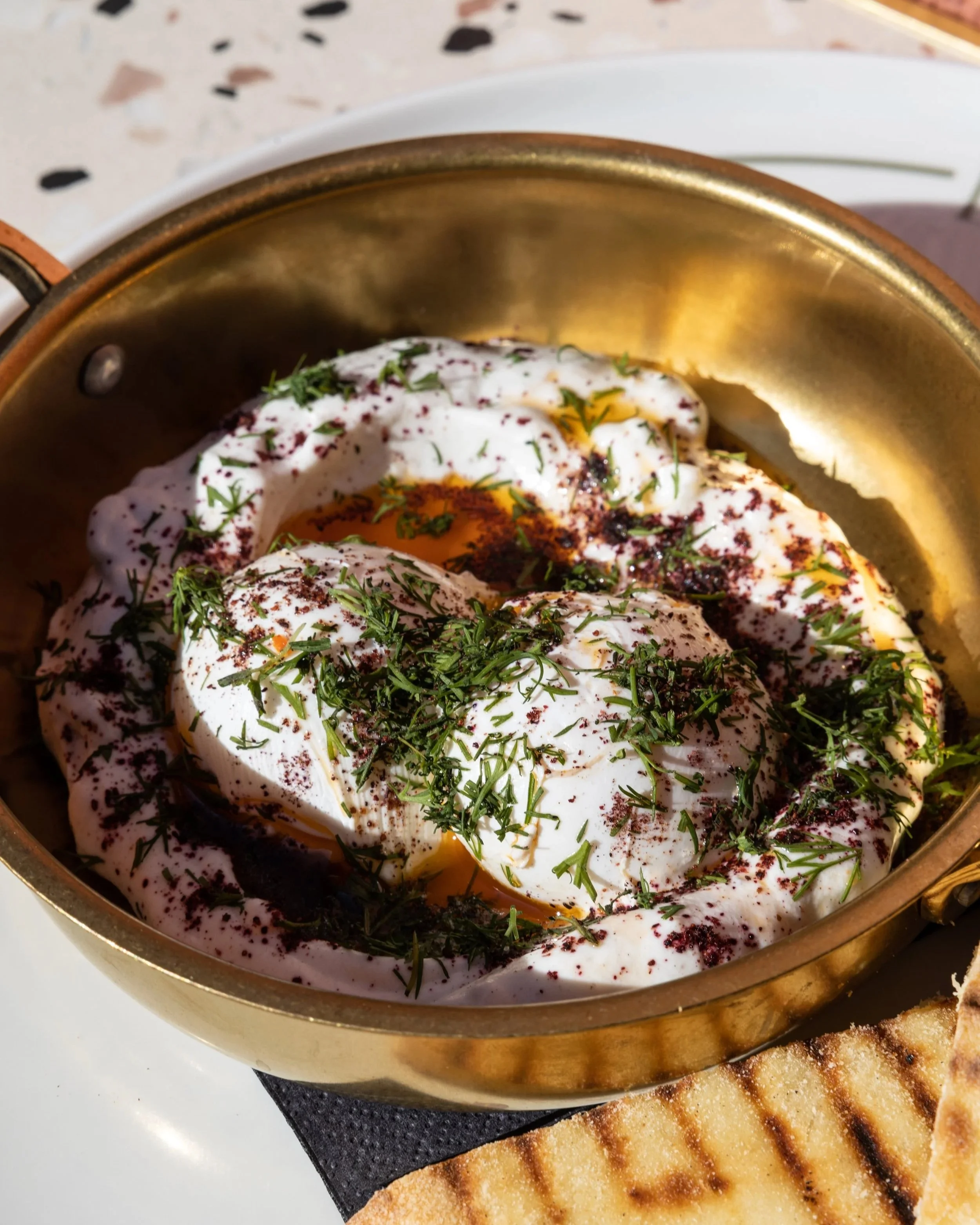Close-up of a cast iron skillet with sunny-side-up eggs garnished with chopped herbs, with grilled bread on a plate in the background.