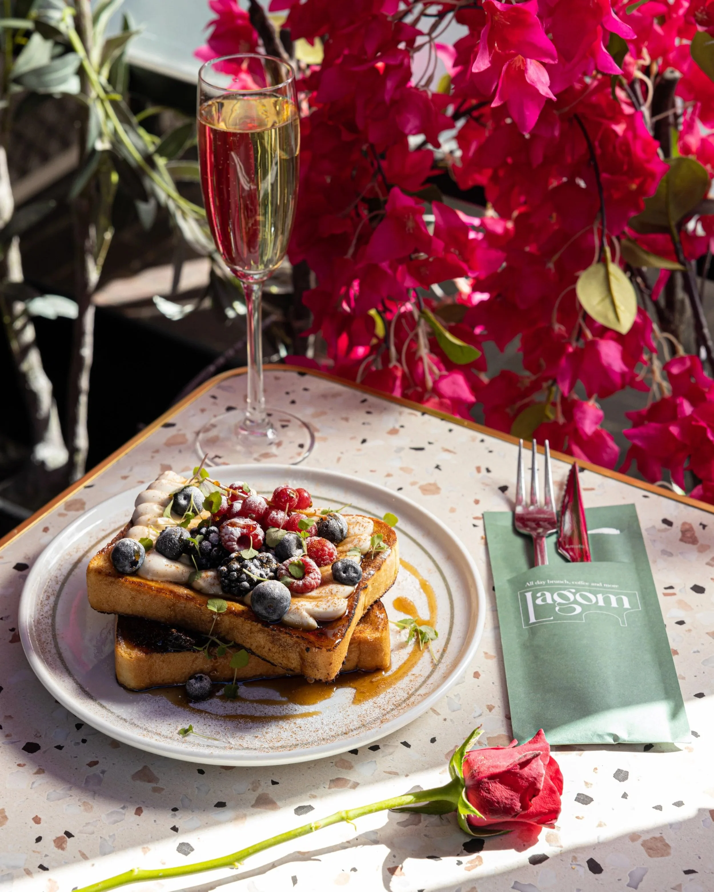 A slice of grilled bread topped with whipped cream, fresh mixed berries, microgreens, and syrup on a white plate. A glass of Champagne and a red rose are on a terrazzo table, with pink bougainvillea flowers in the background.