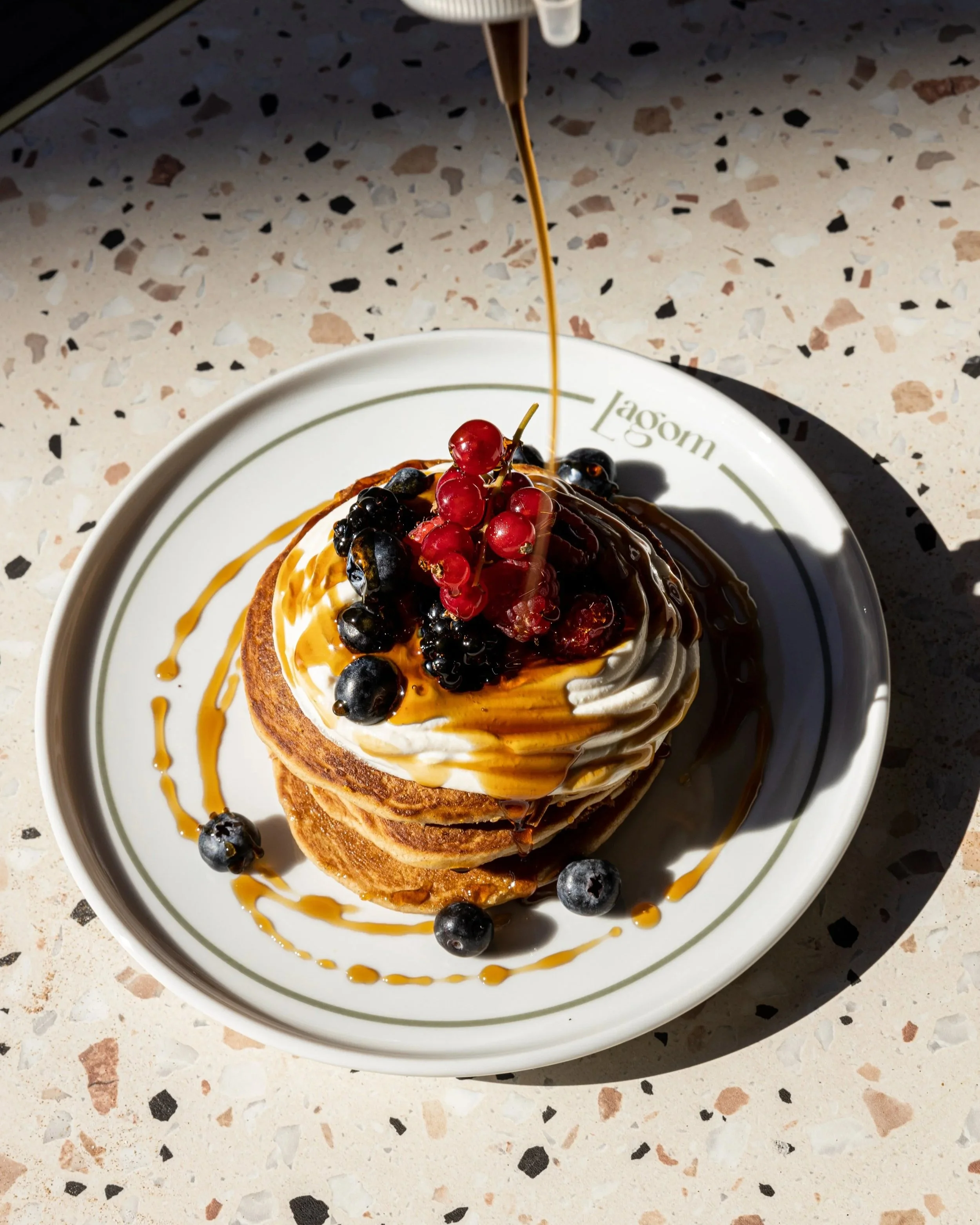 A plate with stacked pancakes topped with whipped cream, red and black berries, caramel syrup, and drizzled with chocolate sauce on a speckled countertop.
