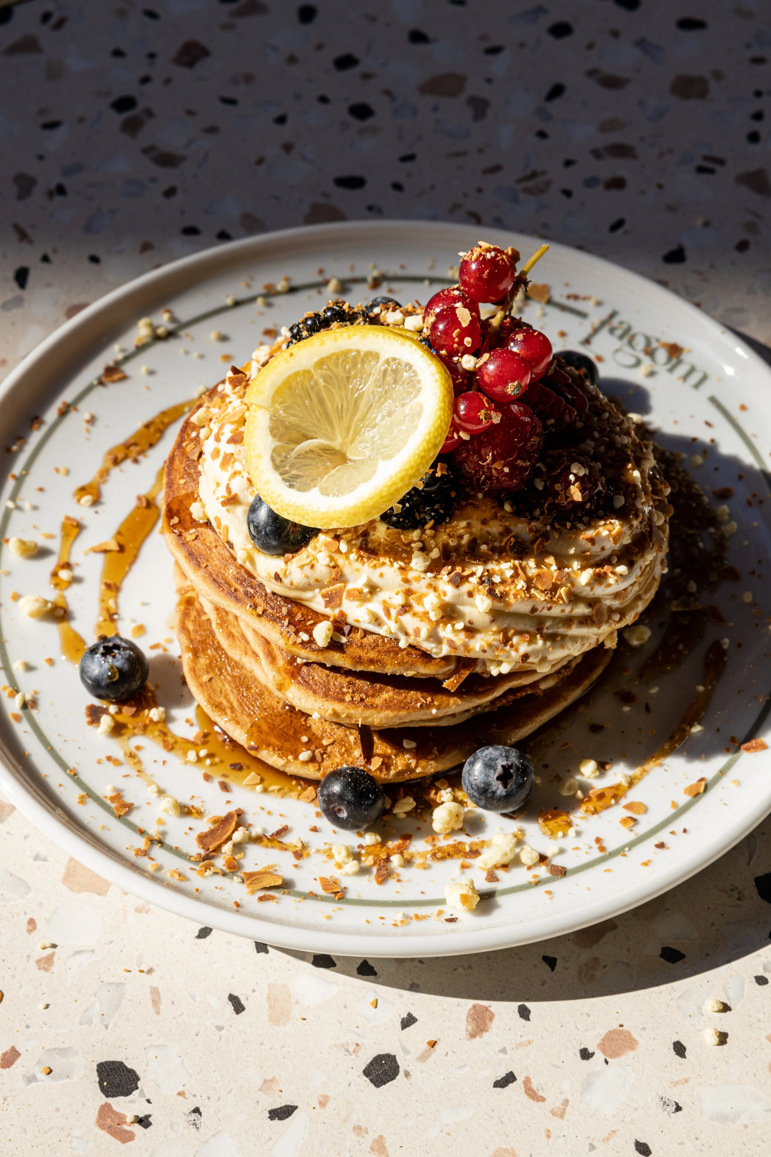 A plate of layered waffles topped with whipped cream, lemon slices, red currants, blueberries, chopped nuts, and caramel sauce.