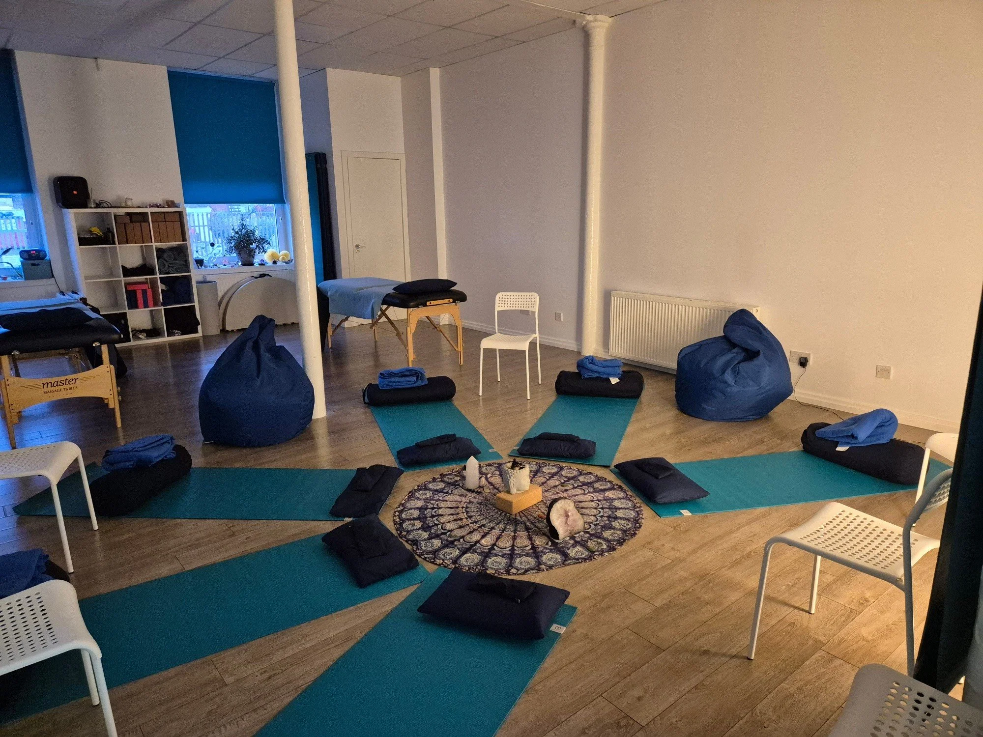 A room set up for a yoga or meditation session with mats, cushions, and blankets on the floor, a small decorative rug with candles and stones, chairs, bean bags, a massage table, and a bookshelf near a window with blinds.