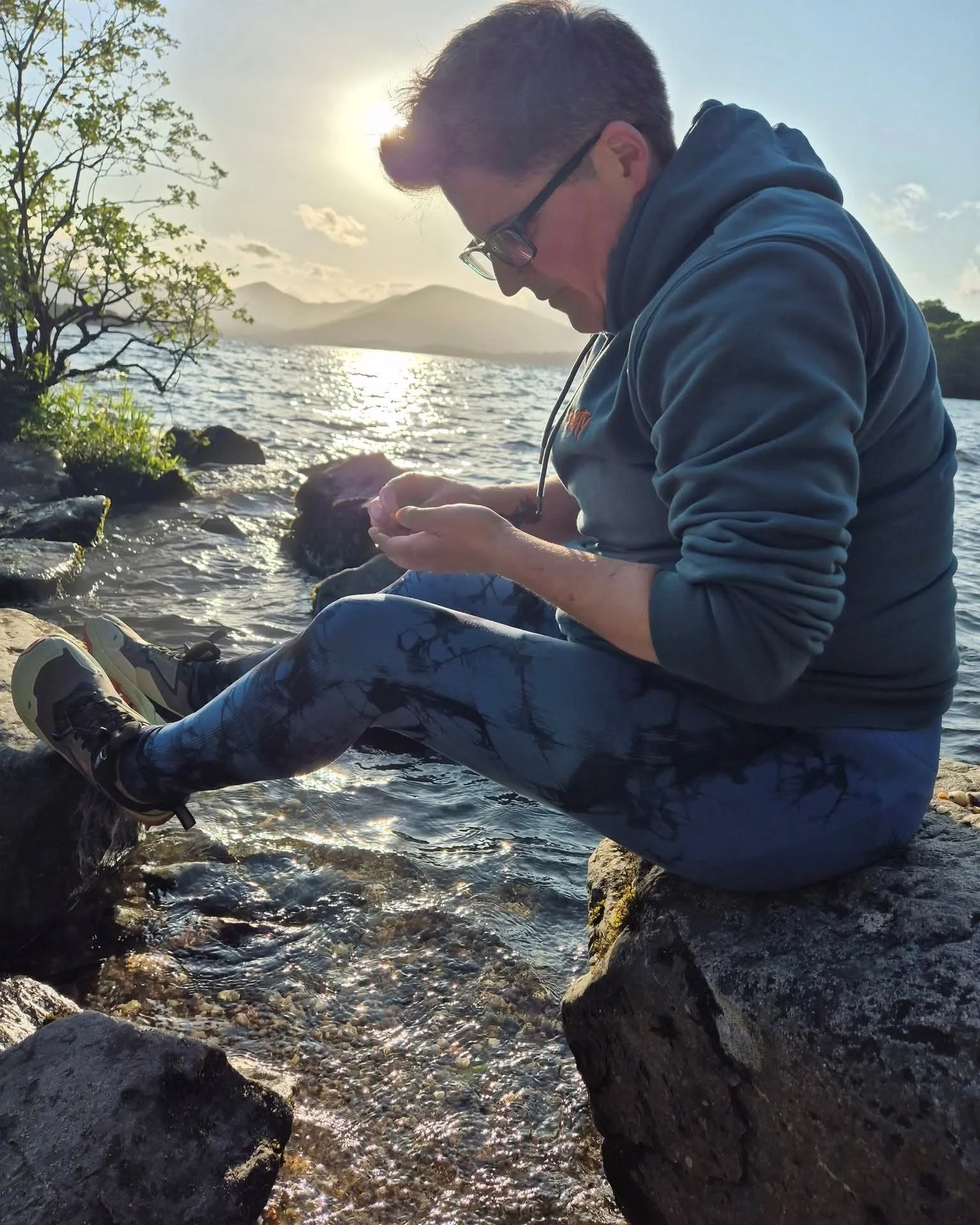 A man with glasses and dark hair, wearing a blue hoodie and leggings, sitting on rocks by a body of water during sunset, looking at his phone.