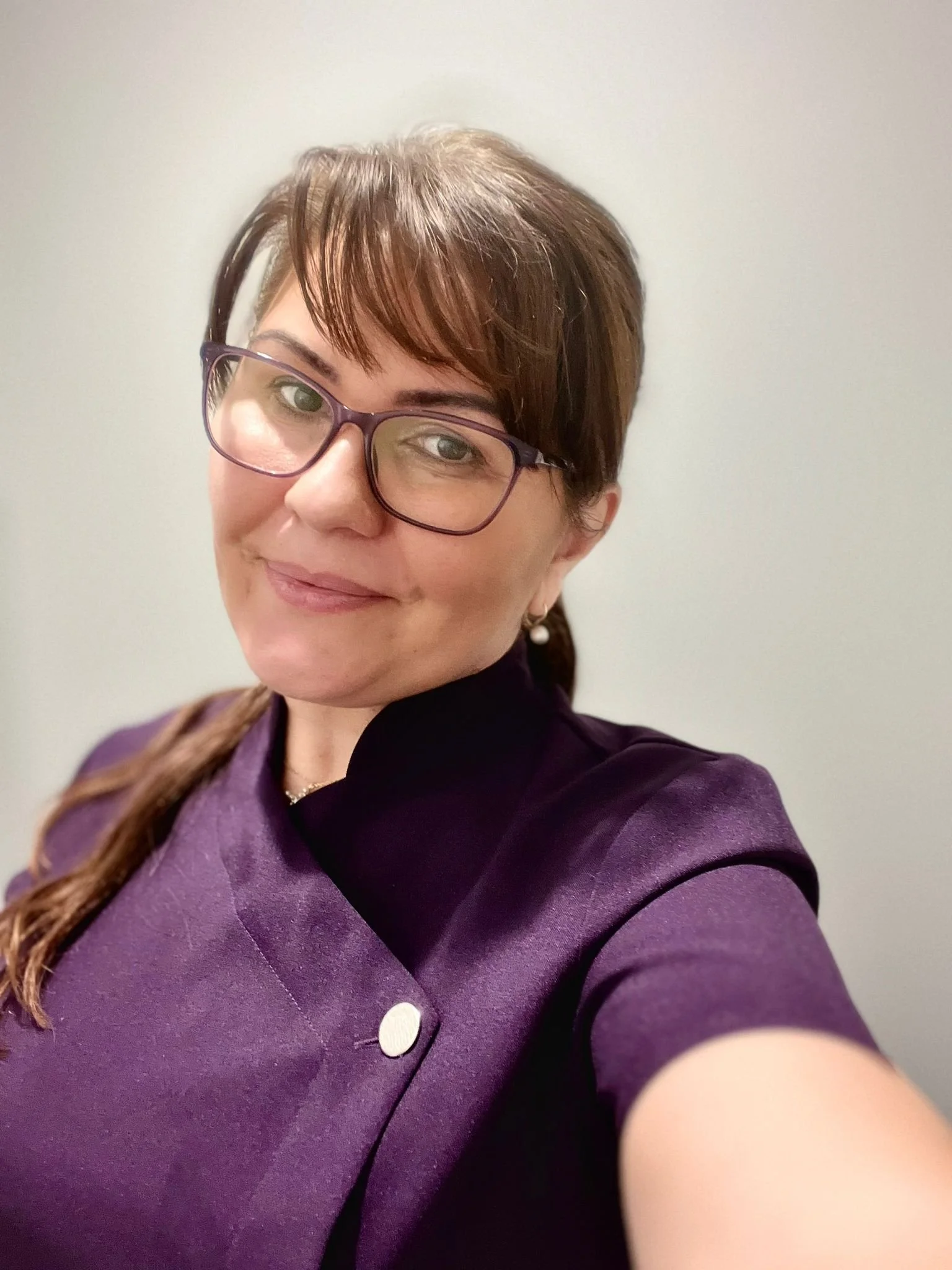 A female therapist with short brown hair wearing glasses and a purple jacket, taking a selfie against a plain off-white wall.