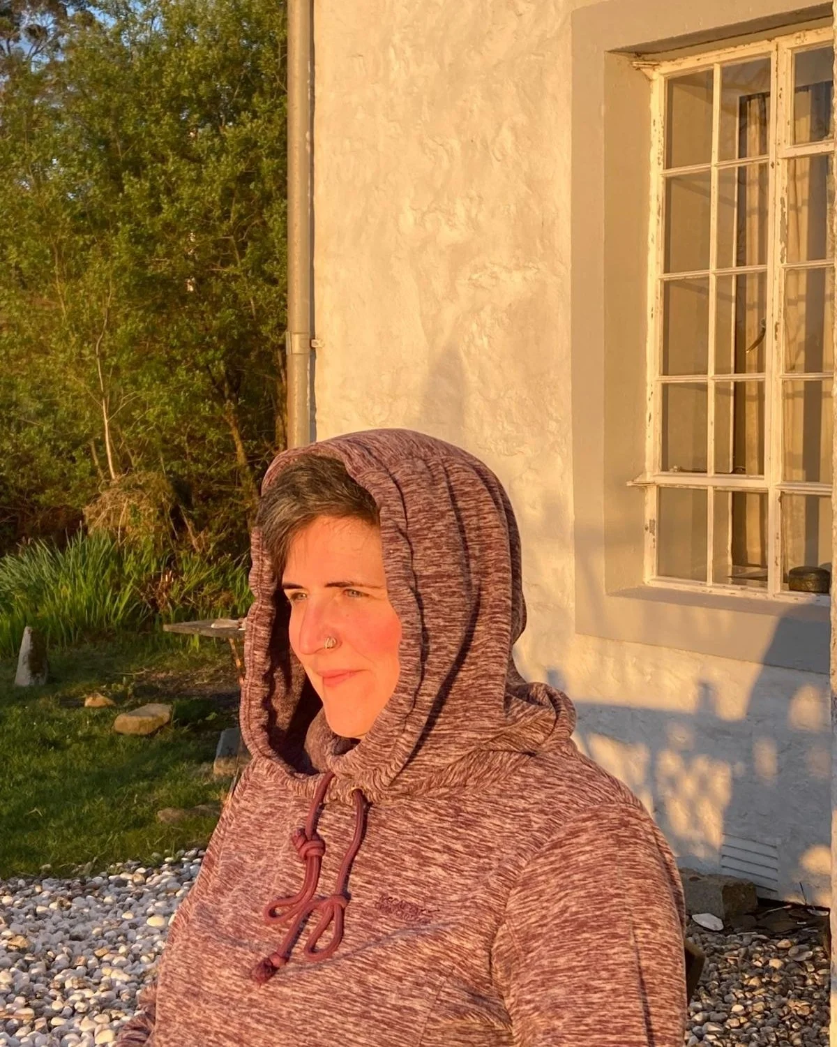 A woman with short dark hair and a nose ring, wearing a maroon hoodie, standing outside on a sunny day, with a white building, green trees, and a small garden in the background.