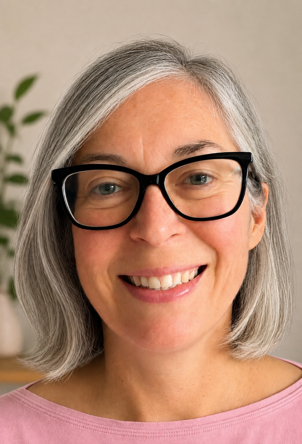 Close-up of a smiling mature woman with gray hair and glasses, wearing a pink top, in an indoor setting with a blurred plant in the background.