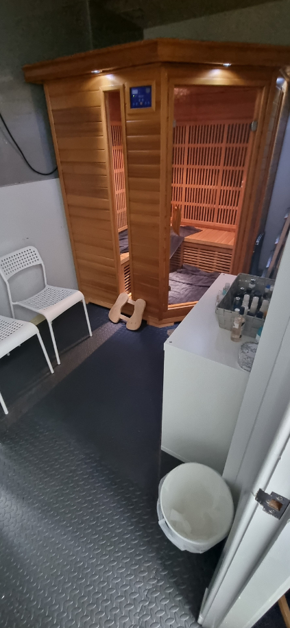 A wooden infrared sauna cabin in a room with plastic chairs, a white cabinet with bottles, and a waste bin.