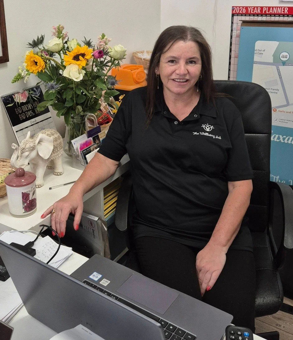 A woman sitting at a desk with a big smile, wearing a black polo shirt with a logo that says "The Welling Hub." The desk has a laptop, a notepad, and various office supplies. Behind her are a vase of colorful flowers, a decorative elephant, and a 2026 planner on the wall.