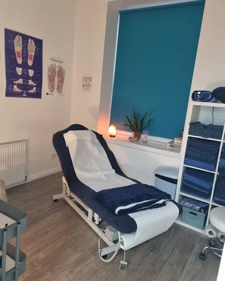 Medical examination room with a reclining chair covered with a white sheet and navy blue towels, a small table, a potted plant, a candle, a medical poster on foot reflexology, shelves with folded towels, a candle, a salt lamp, a machine on the shelf, and a blue window with blinds.
