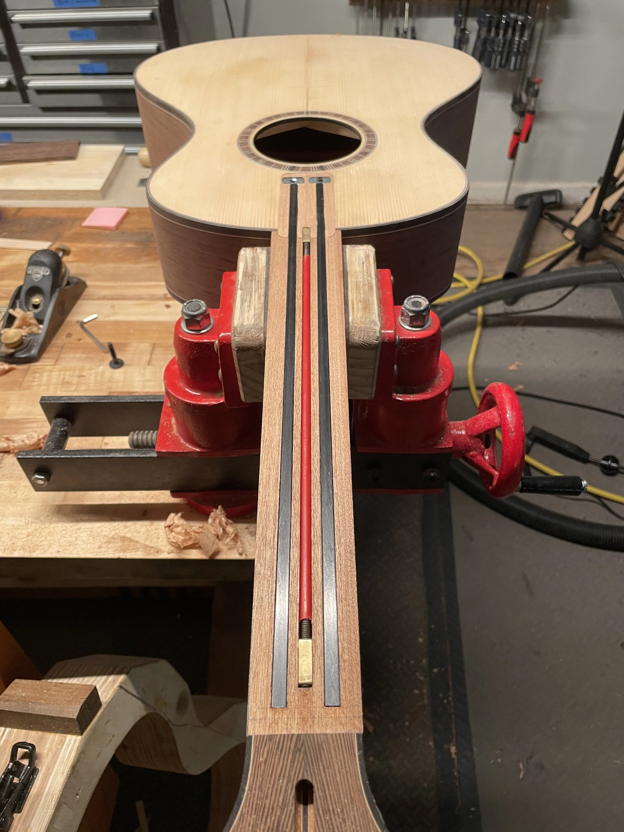 Acoustic guitar neck construction on a workbench with woodworking tools, showing truss rod installation and clamped in vise.