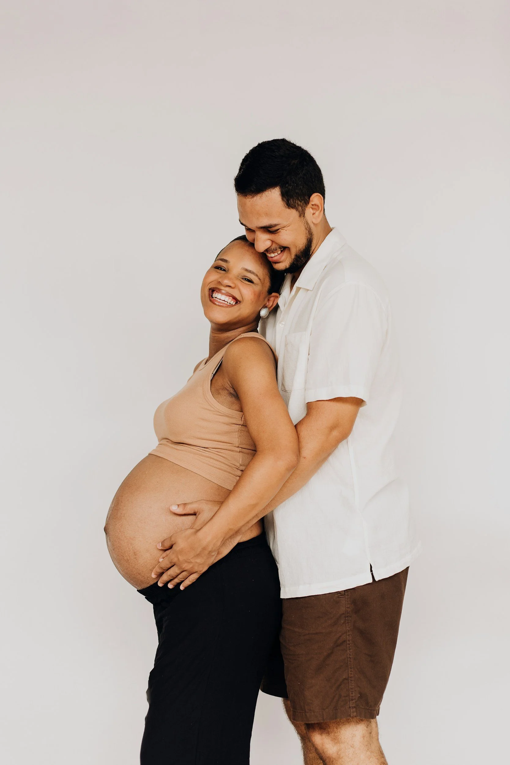A joyful pregnant woman in a nude top and black pants, smiling, being embraced from behind by a man in a white shirt and brown shorts. They are standing against a plain white background, sharing a happy moment.