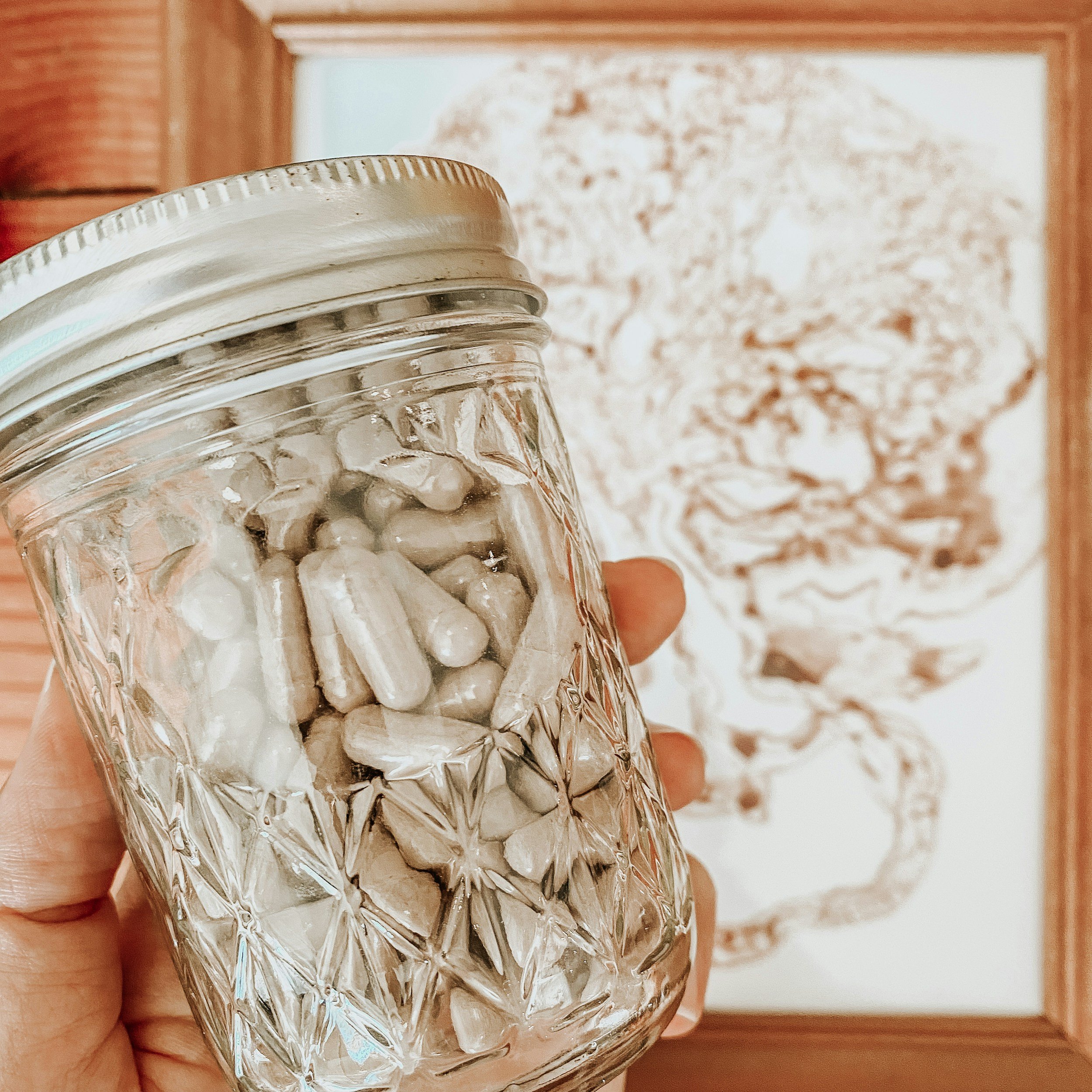 Close-up of a clear textured glass jar with a metal screw-on lid containing encapsulated placenta pills, held by a hand against a background of a placenta print on white paper.