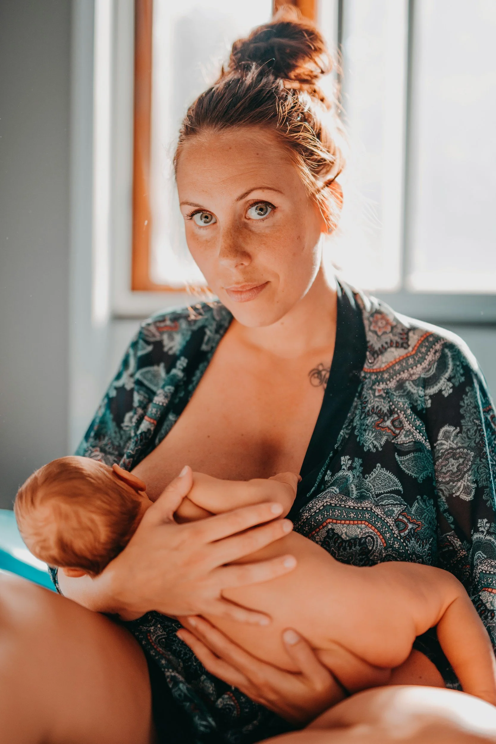 A woman with red hair, blue eyes, and freckles is breastfeeding a baby, sitting near a window with sunlight streaming in.