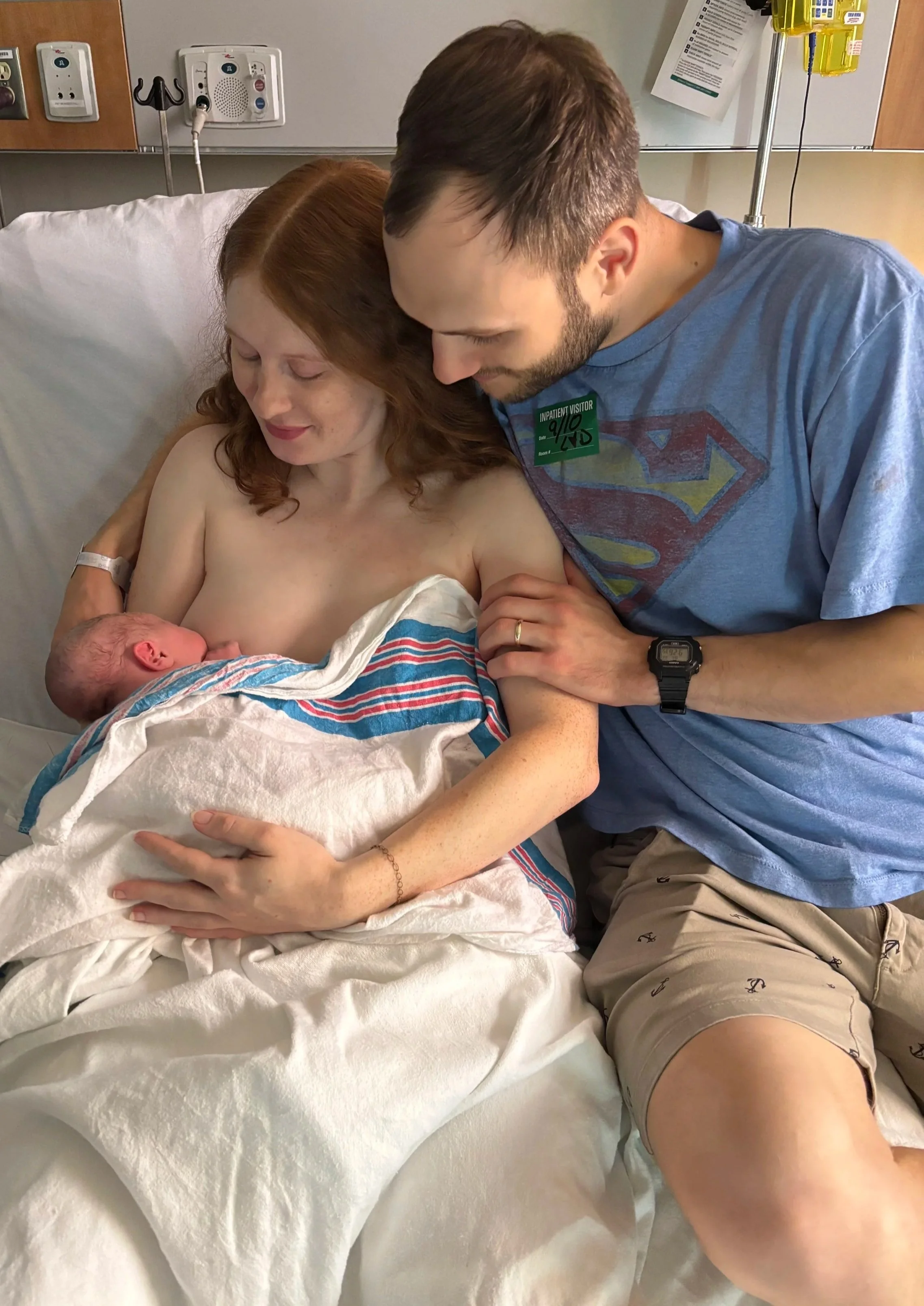 In a hospital room, a woman is sitting in a bed holding her newborn baby. Her husband leans over her, touching her arm and looking happily at the baby.