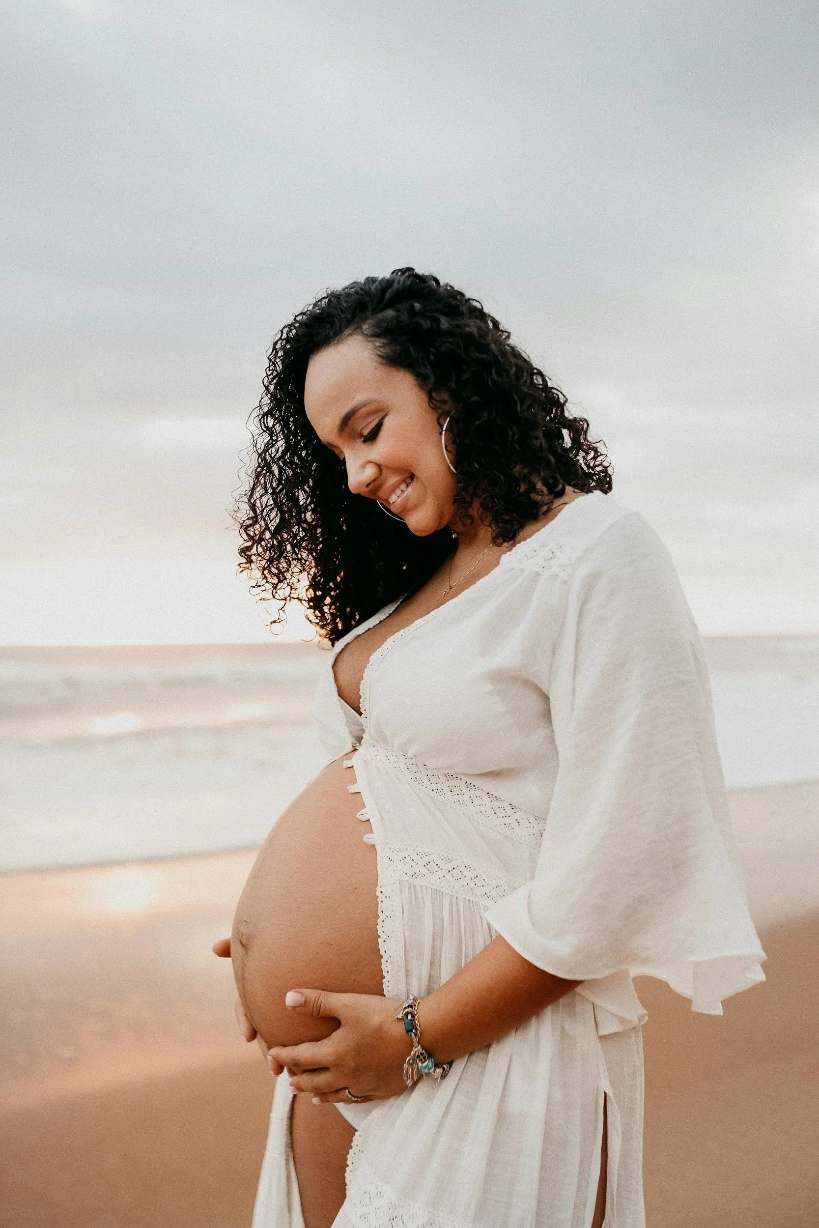 A pregnant woman with curly dark hair smiling while holding her belly, standing on a beach with a cloudy sky in the background.