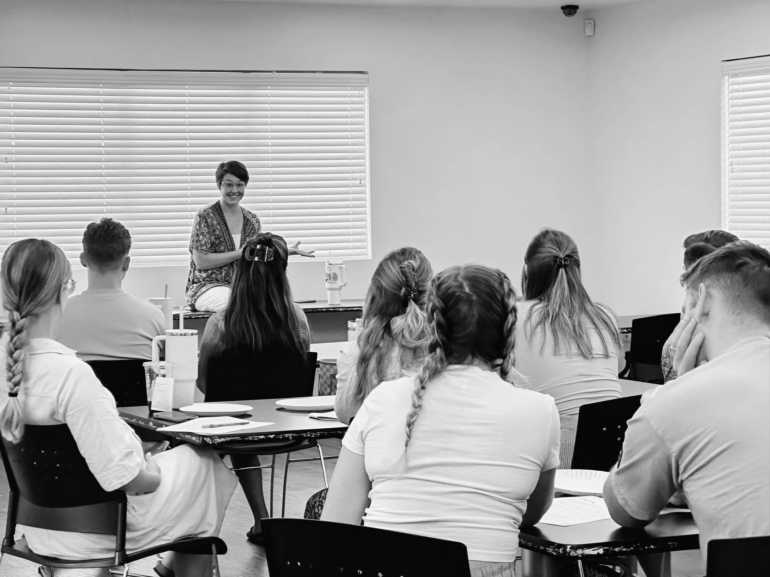 A woman sitting and speaking to a group of students seated at tables in a classroom.