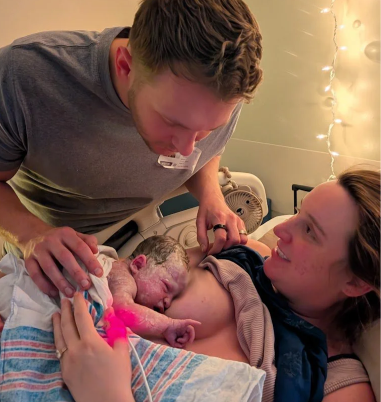 A woman giving birth in a hospital room with an excited expression, and her husband is leaning over her, witnessing the birth of a newborn baby, surrounded by warm lighting.