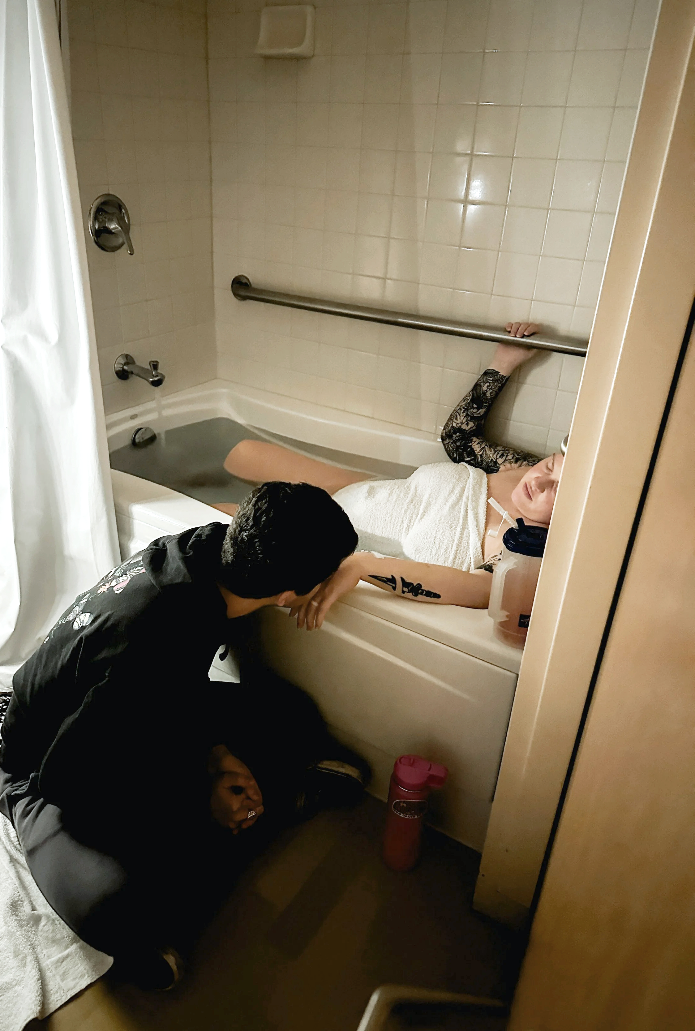 A woman in labor is resting between contractions in a bathtub with a towel over her body while her husband kisses her arm that is on the side of the tub.