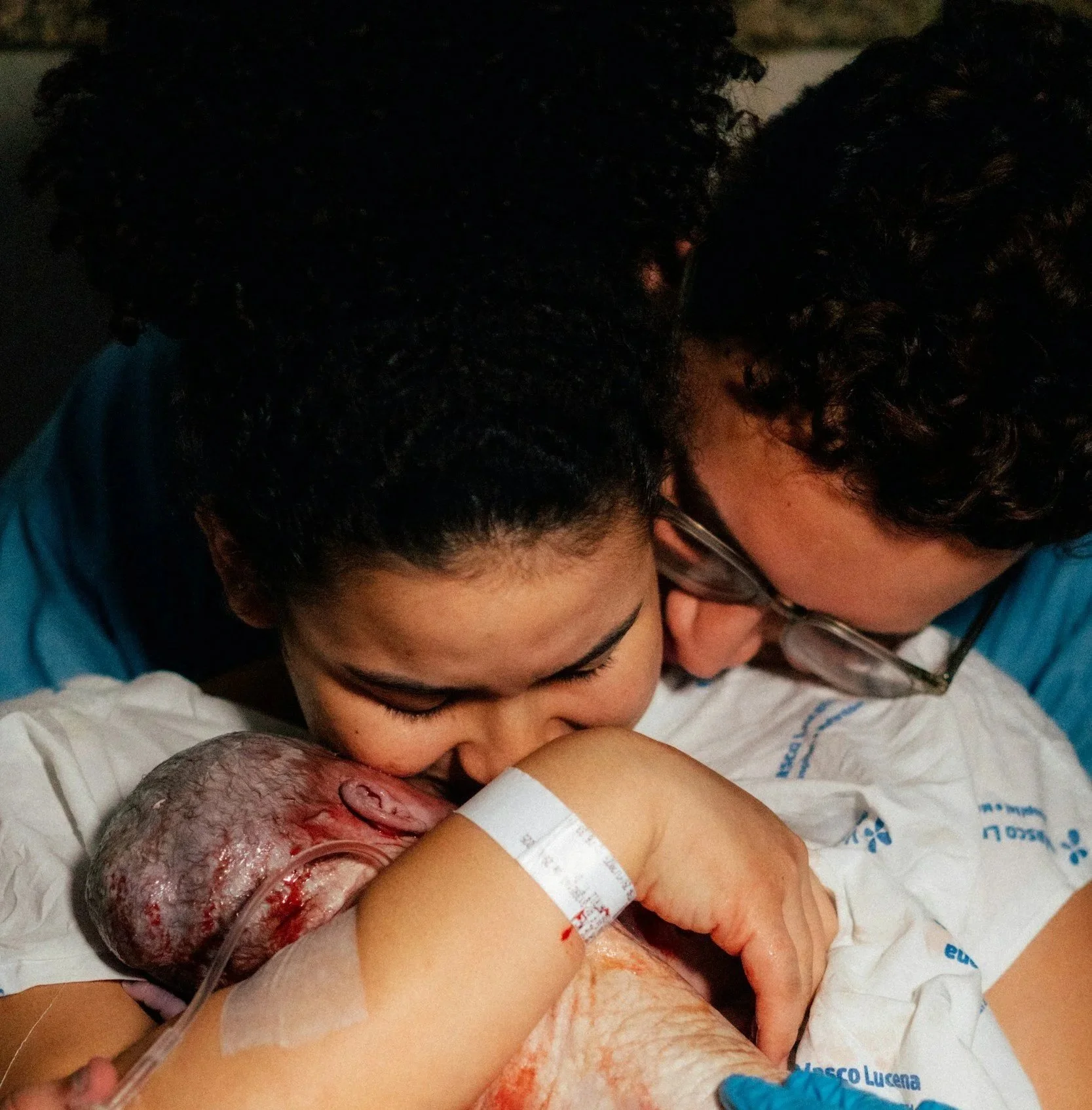 A couple is snuggled close together and holding a newborn baby, who is still attached to the umbilical cord, immediately after birth in a hospital setting.