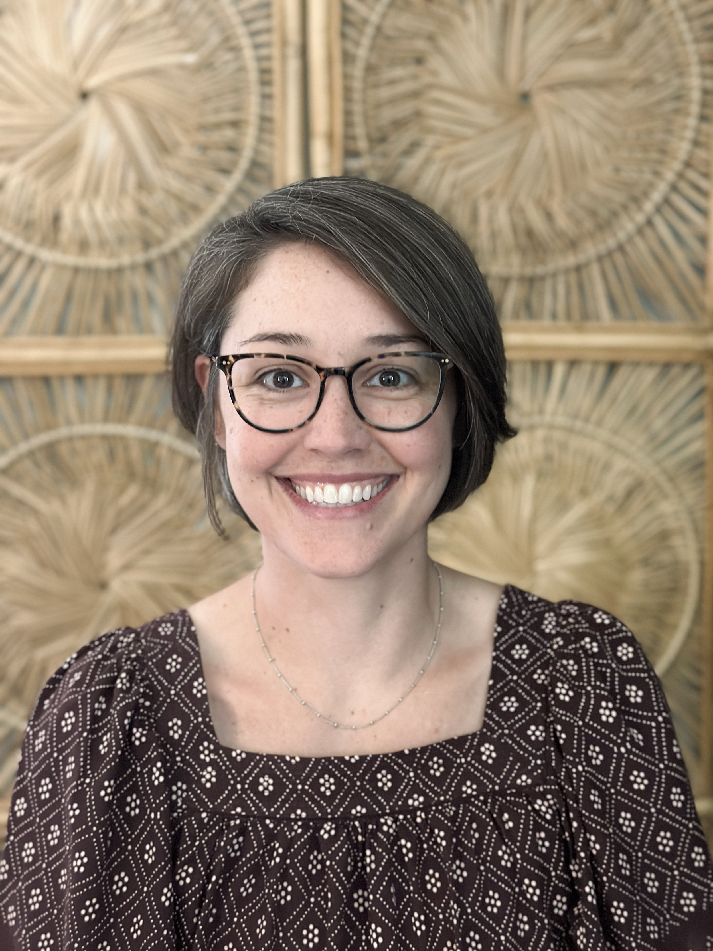 Close-up of a smiling woman with glasses, short dark hair, wearing a patterned top and a delicate necklace, with a decorative woven wall background.
