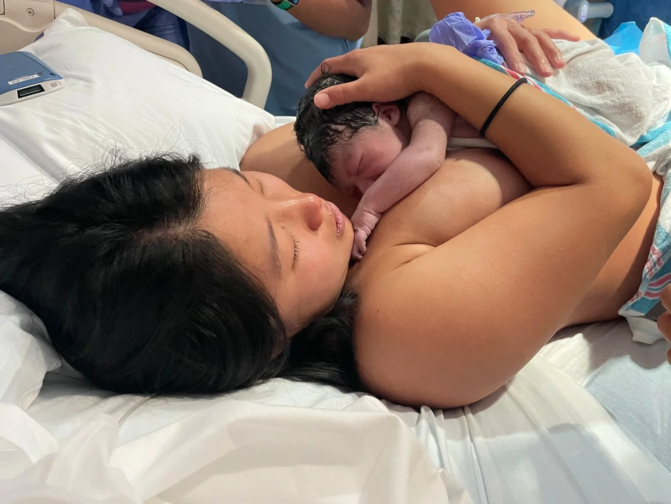 A woman with dark hair lying in a hospital bed holding her newborn baby close to her chest, with the baby’s head resting between her breasts.