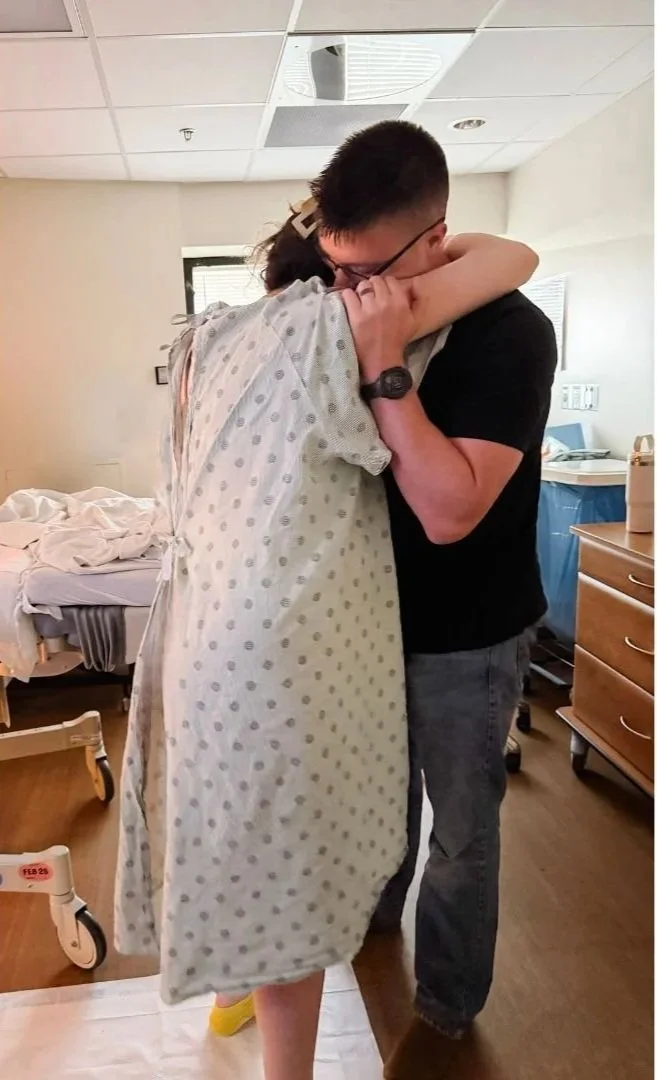 A woman in labor is hugging her husband and leaning on him for support in a hospital room. The woman has her face buried in the man's shoulder.