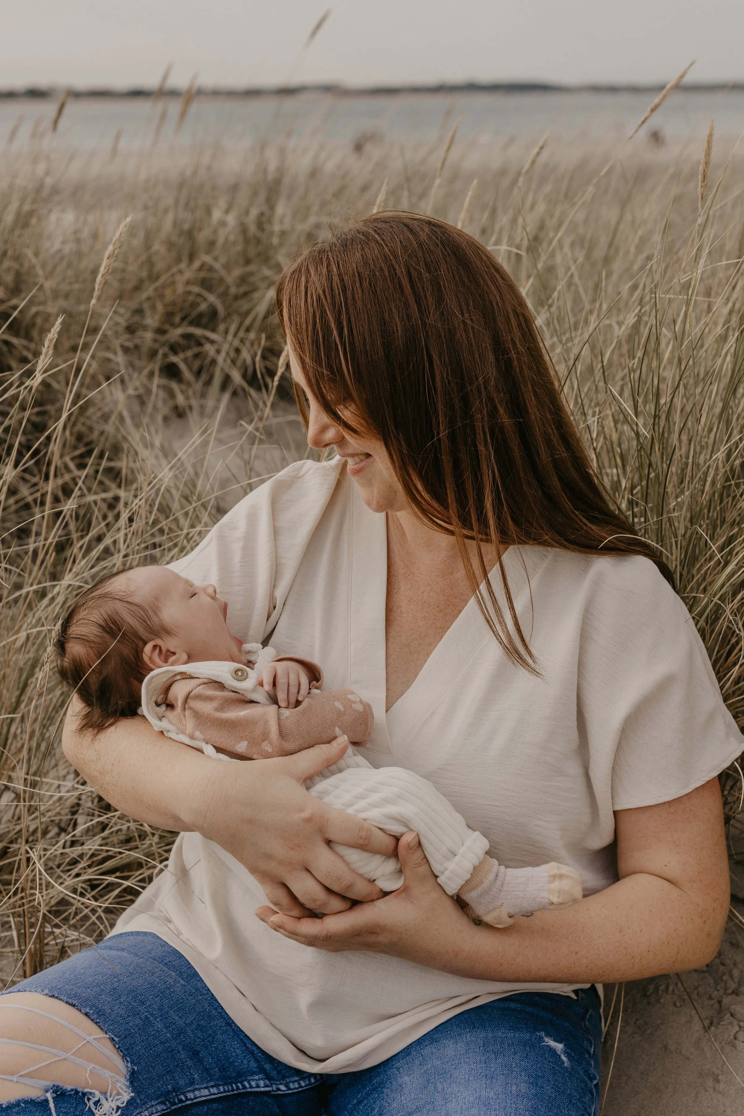 Woman sitting in tall grass by the beach, holding a baby and smiling.