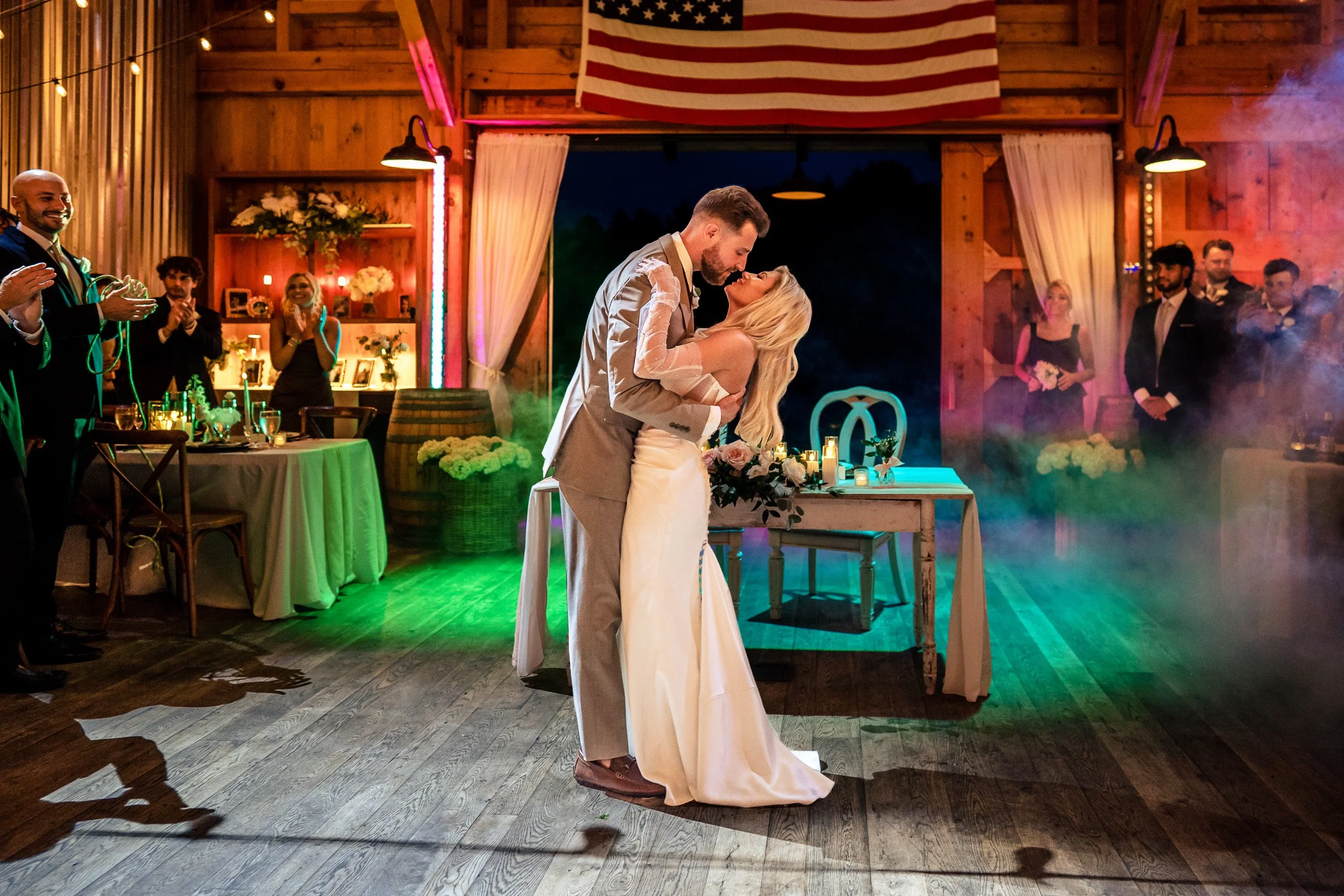 Ava and Jacob kiss on the wooden reception dance floor beneath colored lights.