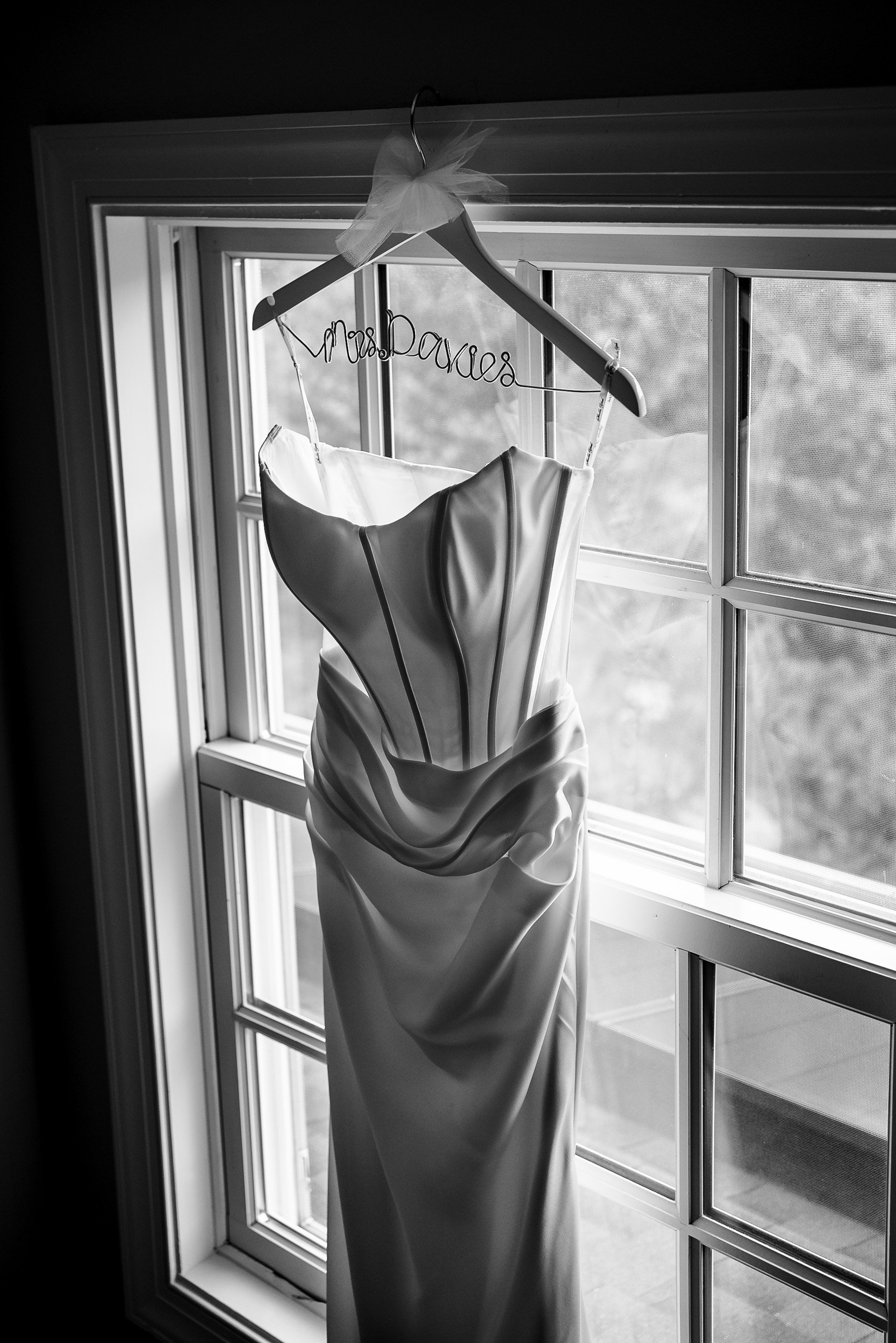 Black-and-white photograph of Ava’s wedding dress hanging beside a window.