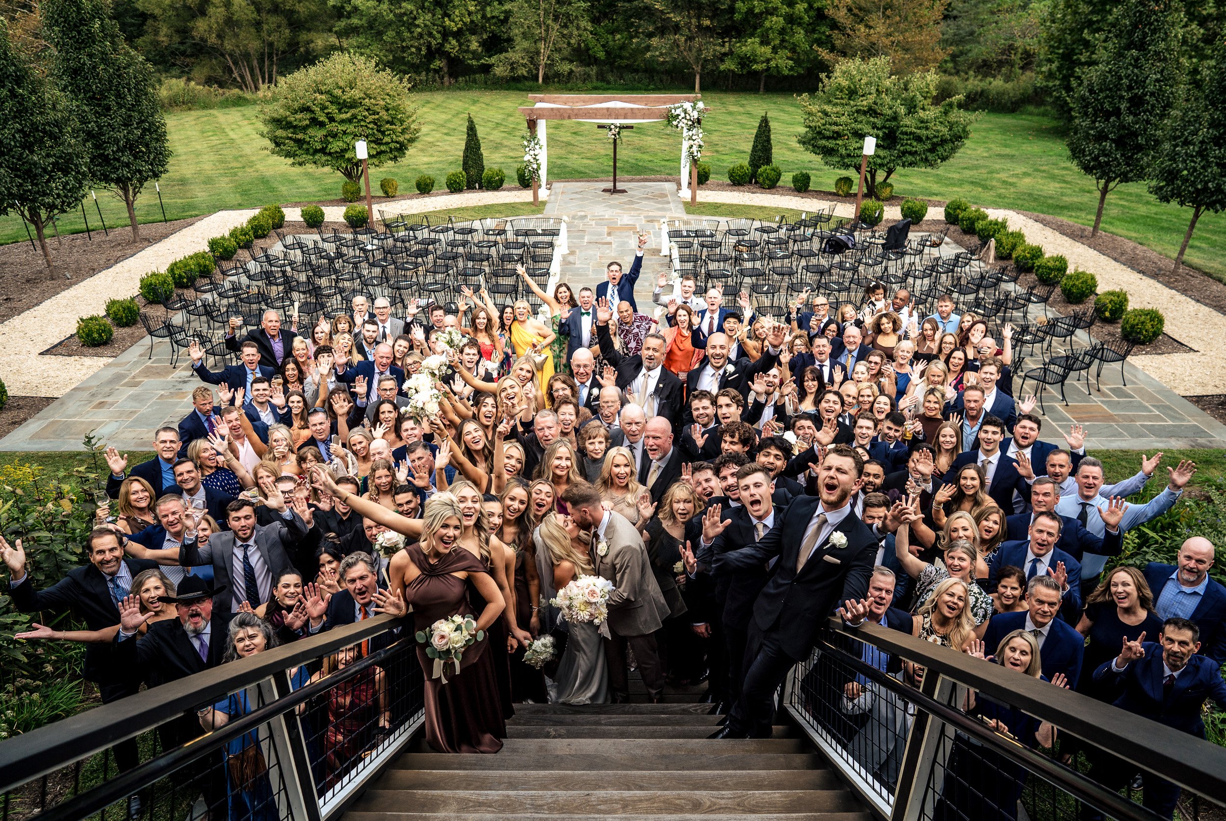 Ava and Jacob gather with their wedding guests for a large outdoor group photograph beside the ceremony seating.