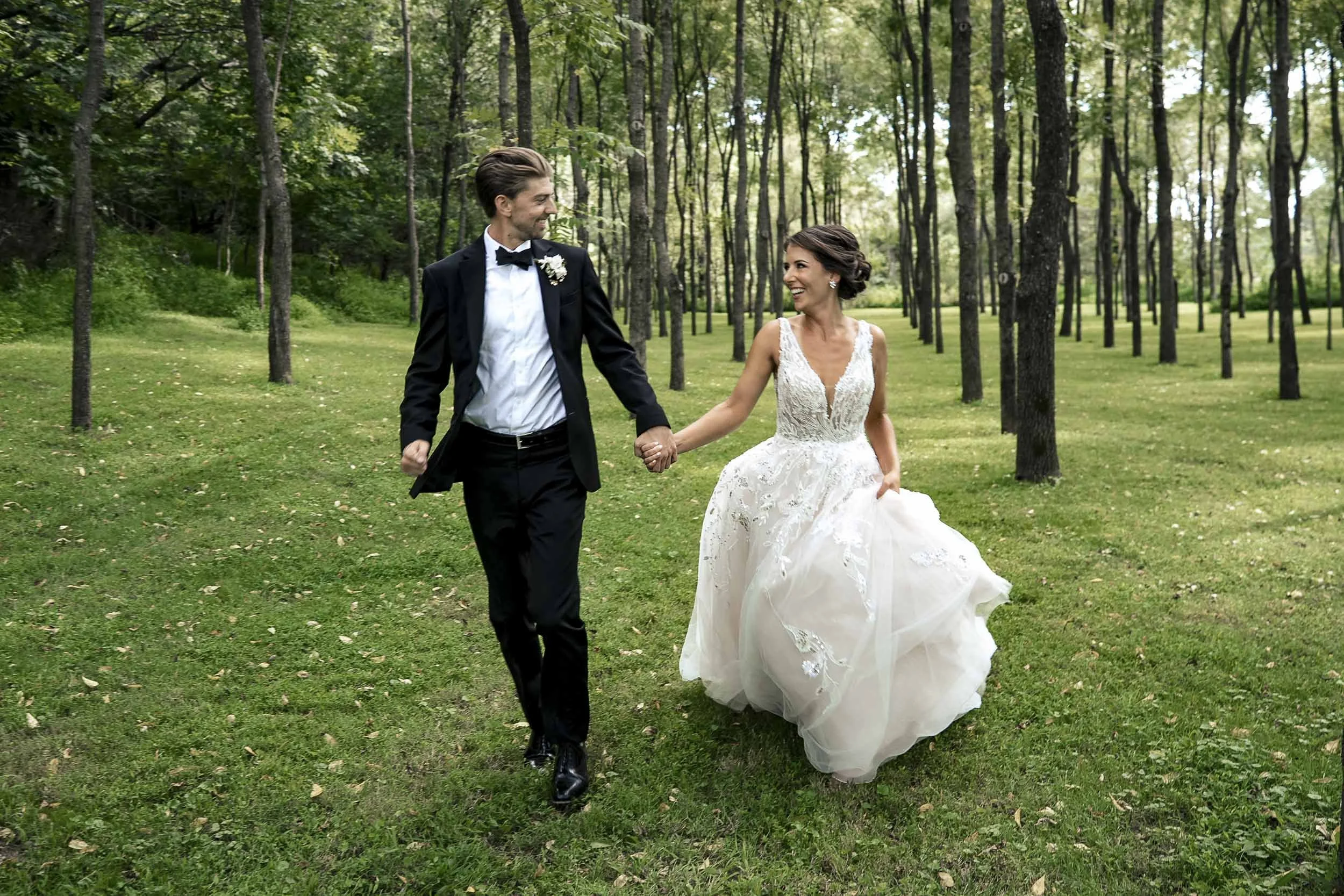 Sarah and Andrew holding hands and smiling at each other as they walk through a grove of trees.