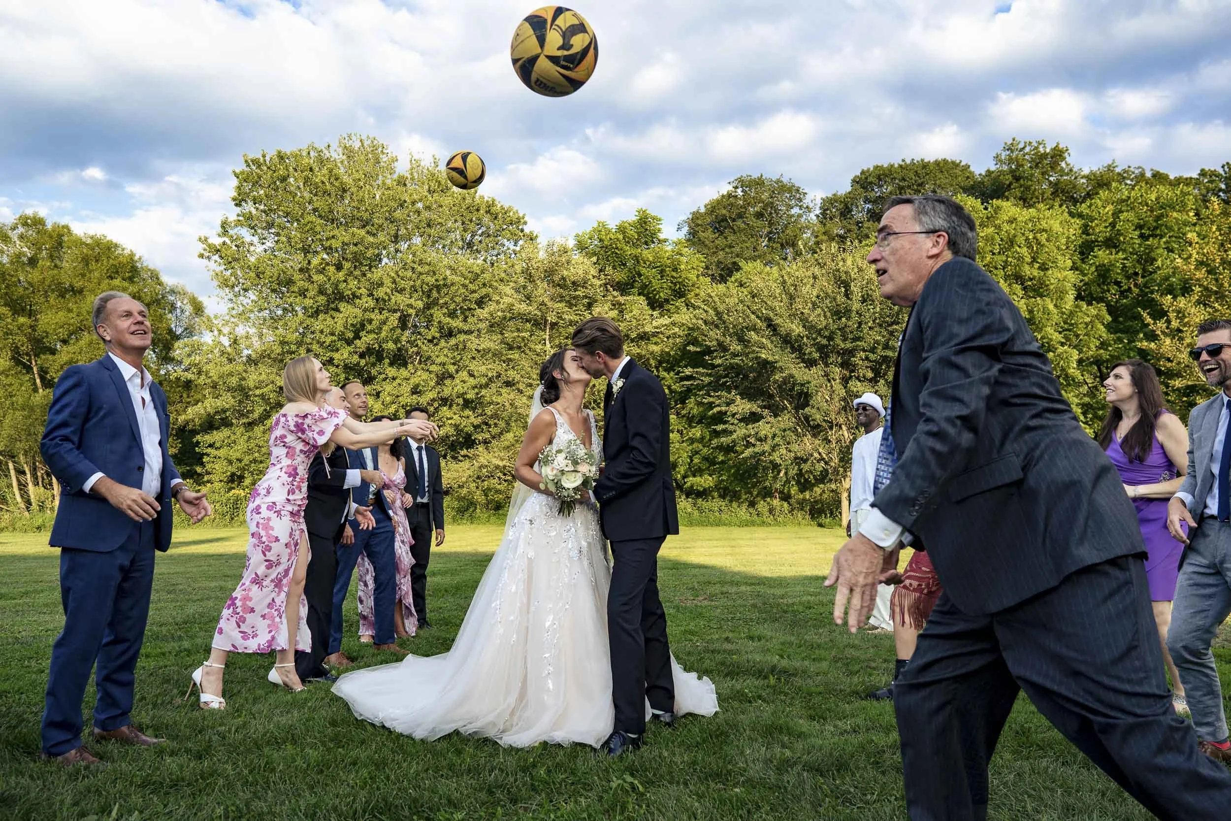 Sarah and Andrew kissing on a lawn while members of their volleyball club pepper volleyballs around them.