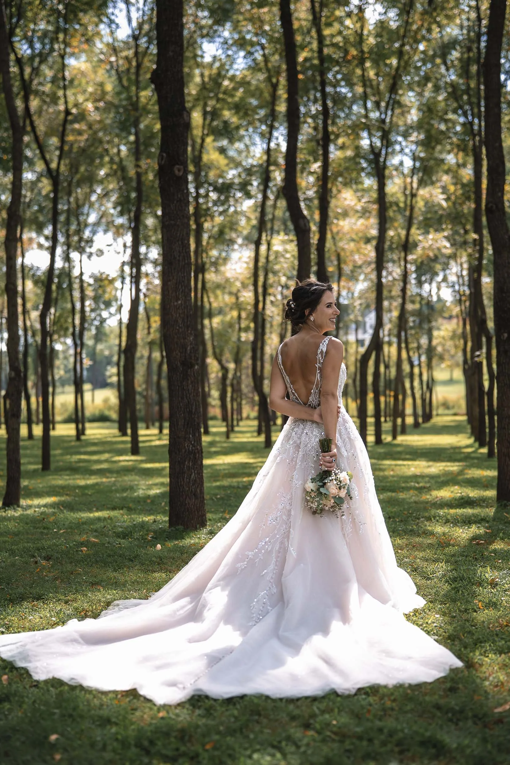 Sarah looking over her shoulder among tall trees, with her wedding dress train spread across the grass.