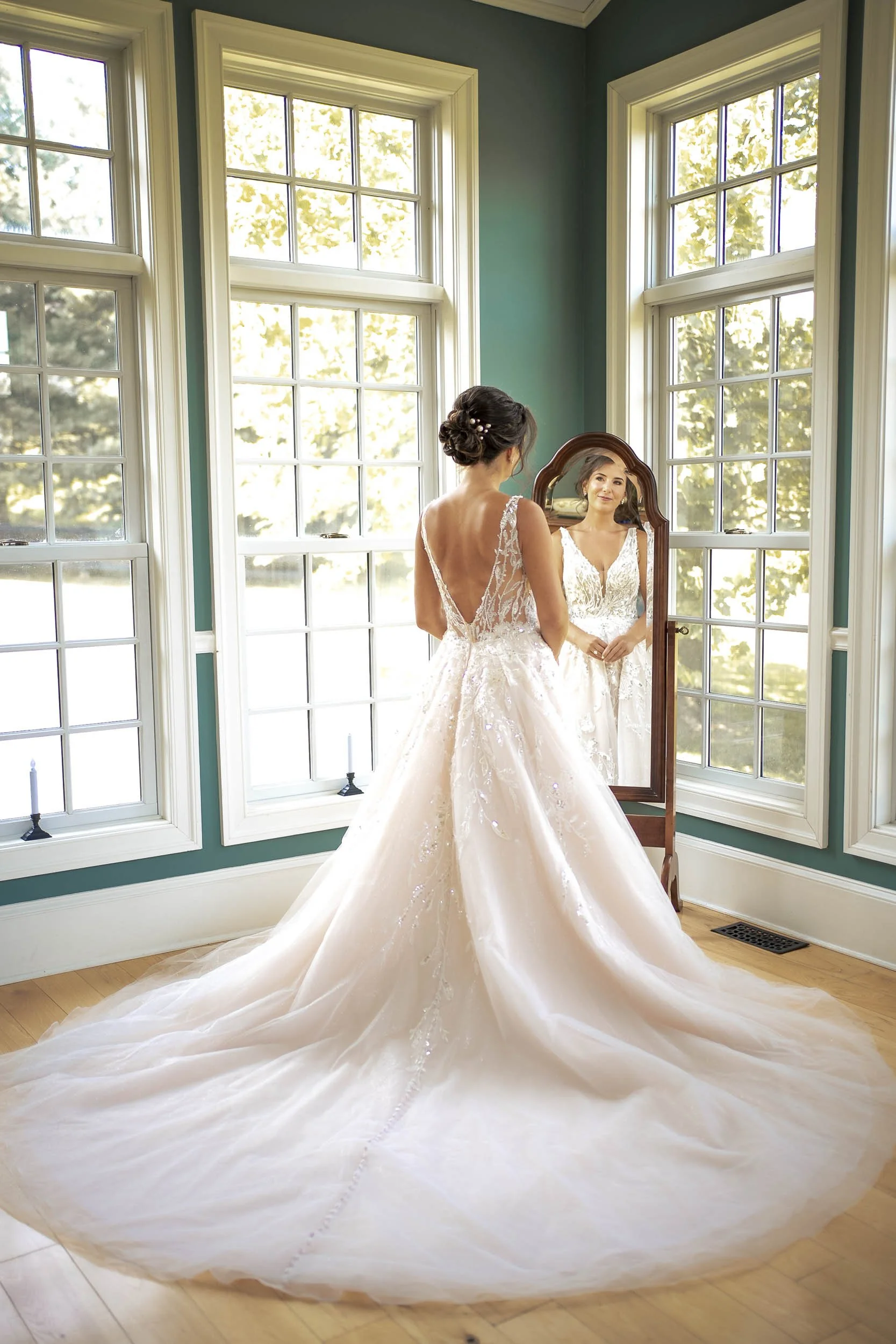 Sarah looking into a standing mirror with her lace wedding gown and long train spread across the sunlit floor.