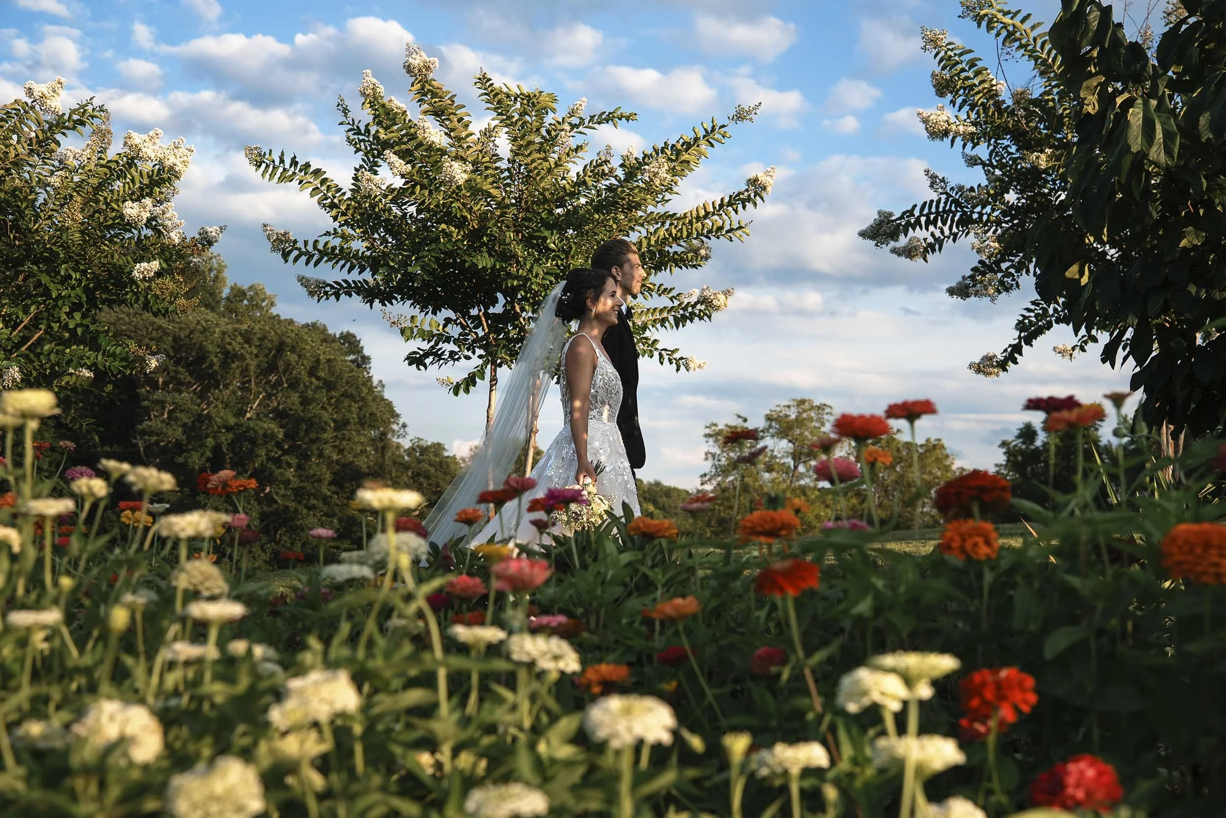 Sarah standing in a garden, framed by colorful flowers and leafy branches beneath a blue sky.