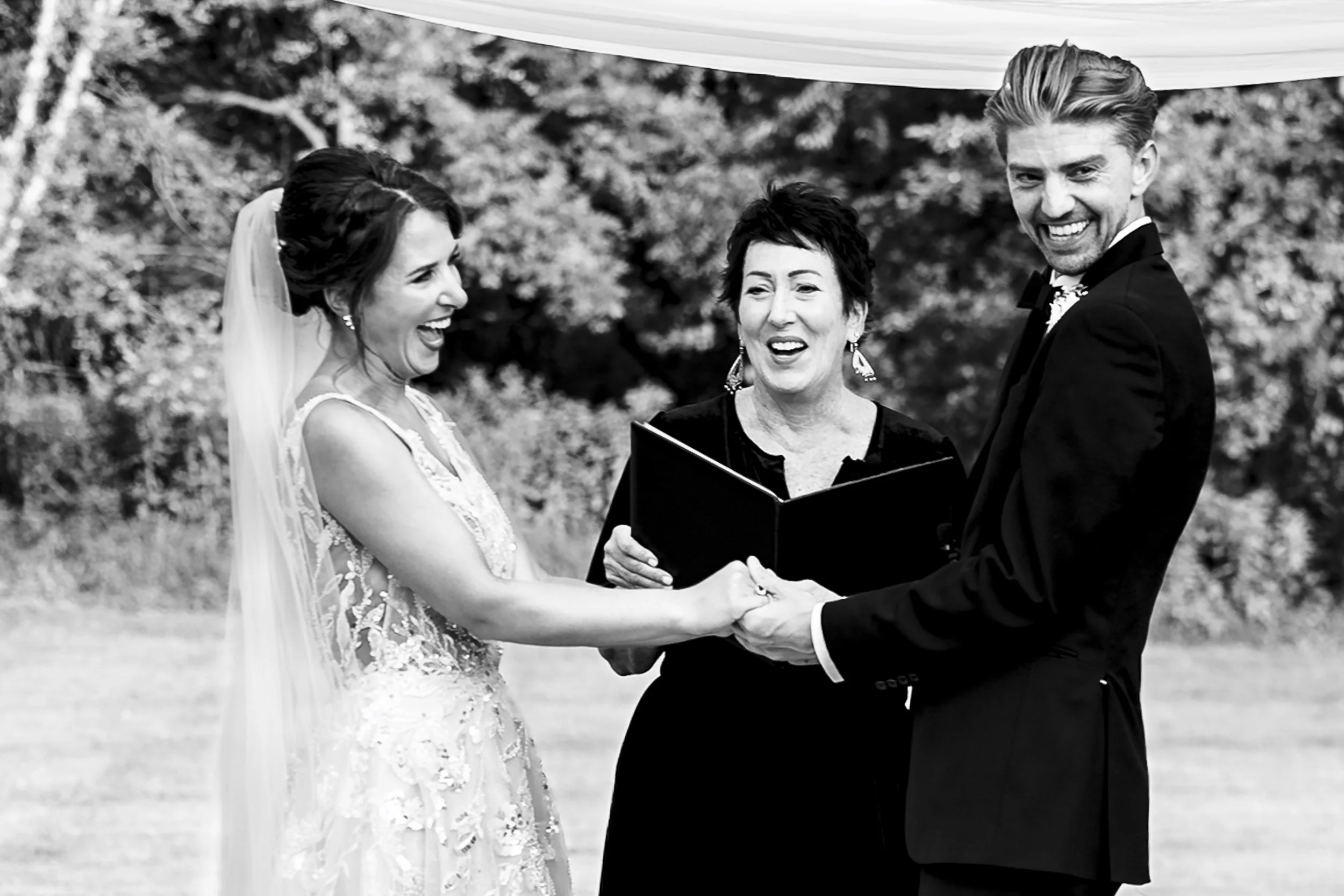 Black and white photograph of Sarah and Andrew holding hands and laughing with their wedding officiant.