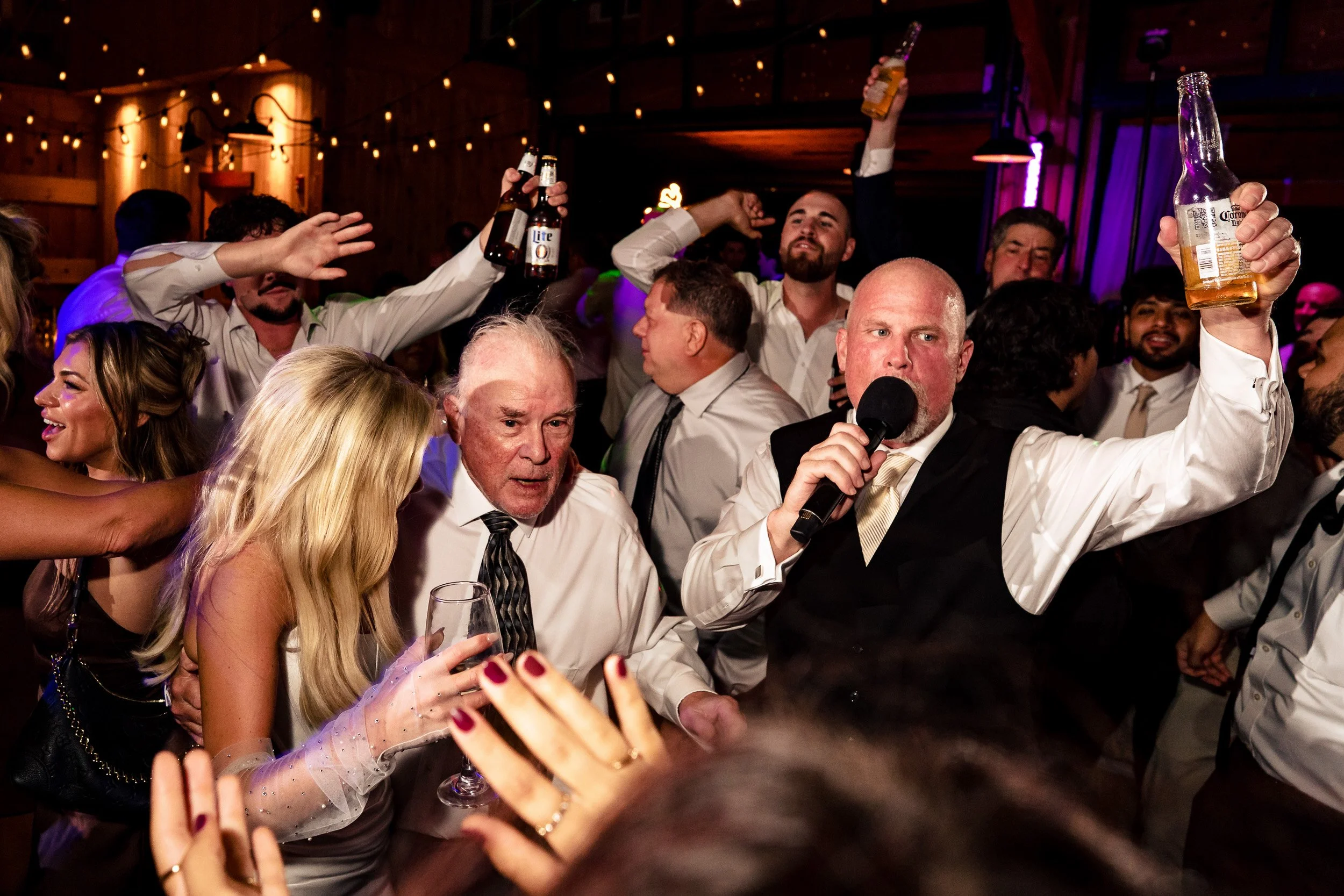 Ava's dad holds a microphone and raises a bottle while Ava and other guests celebrate around him.