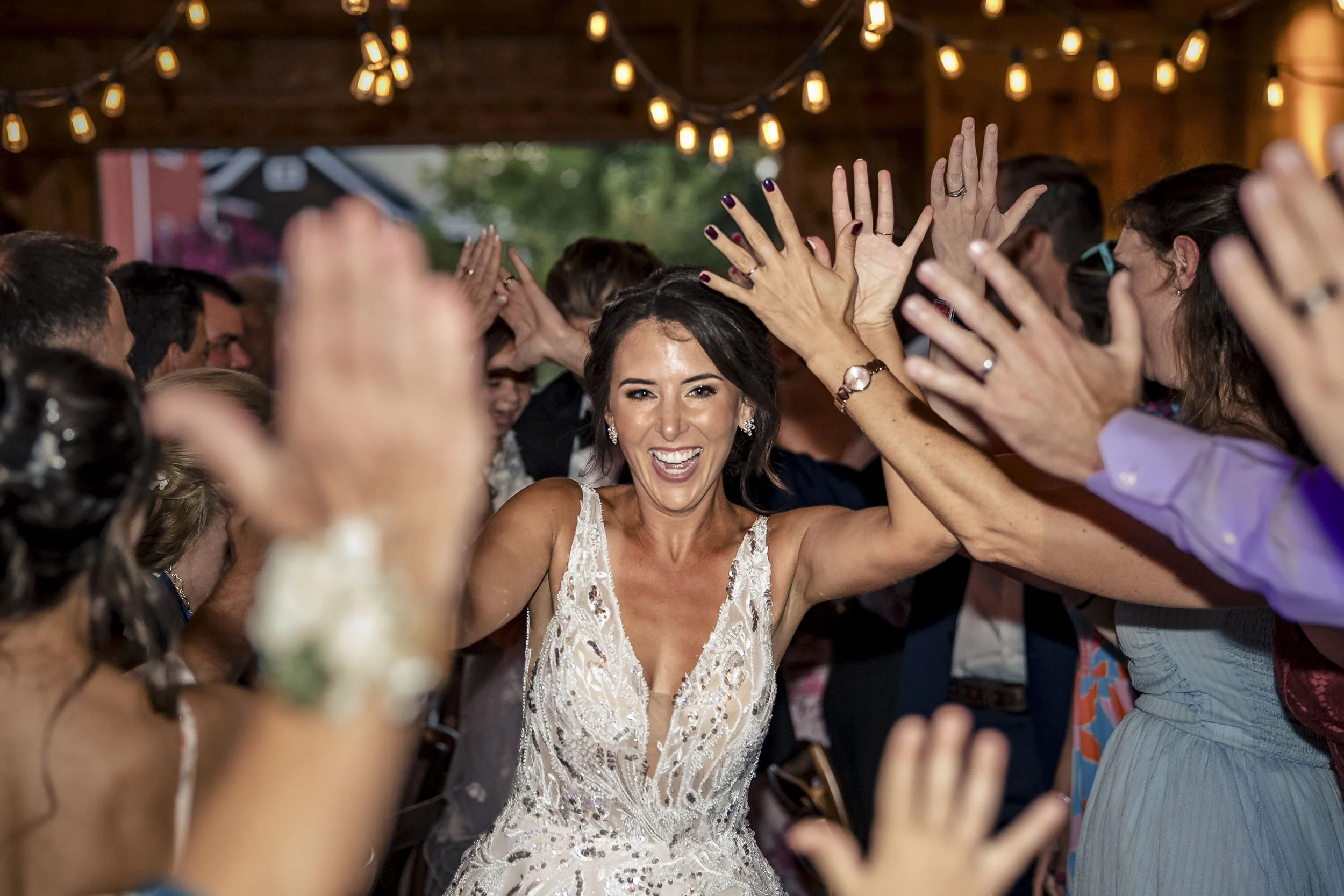 Sarah smiling as she passes between guests with their hands raised beneath reception string lights.