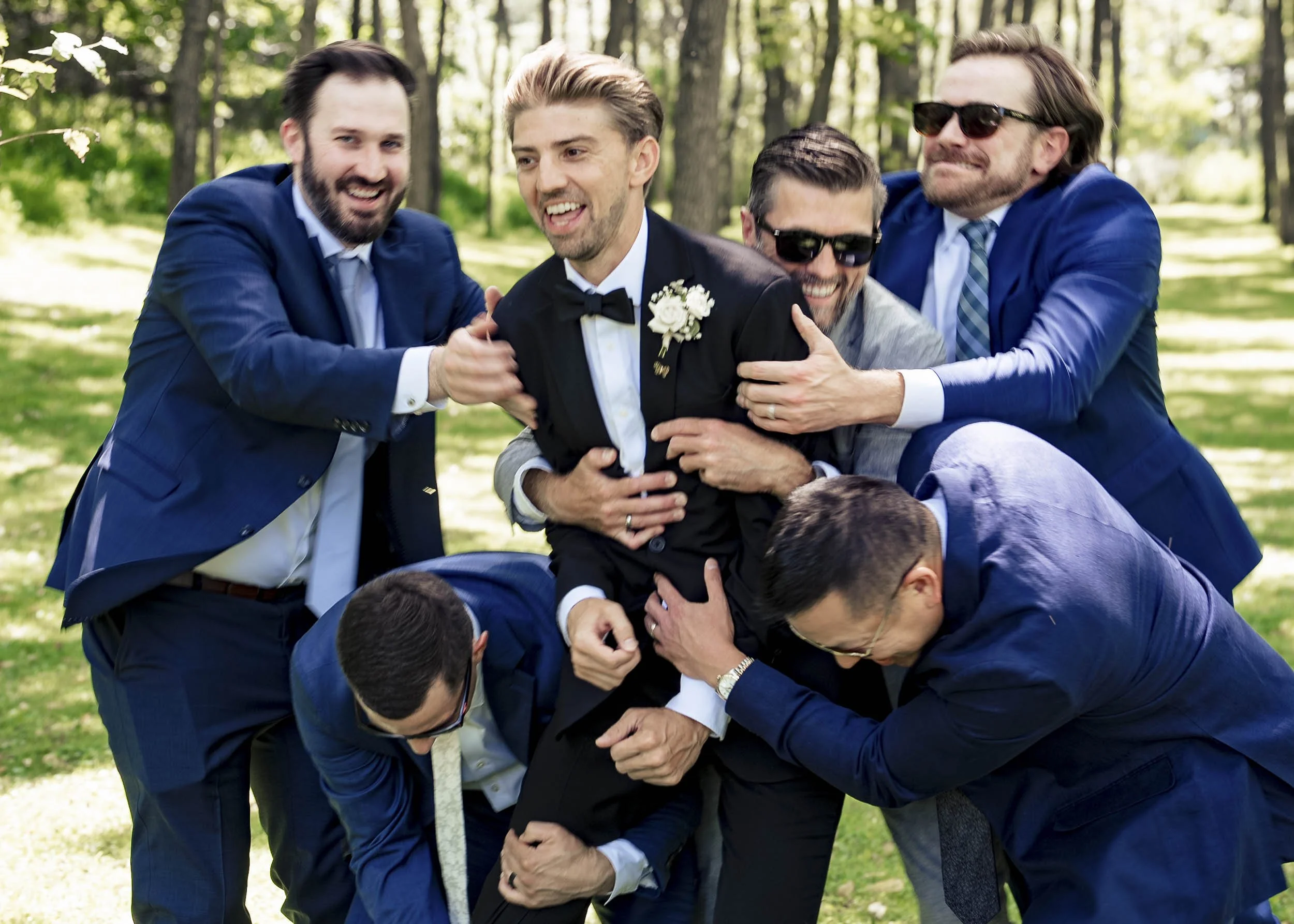 Andrew laughing as his groomsmen in blue suits crowd around him beneath the trees.