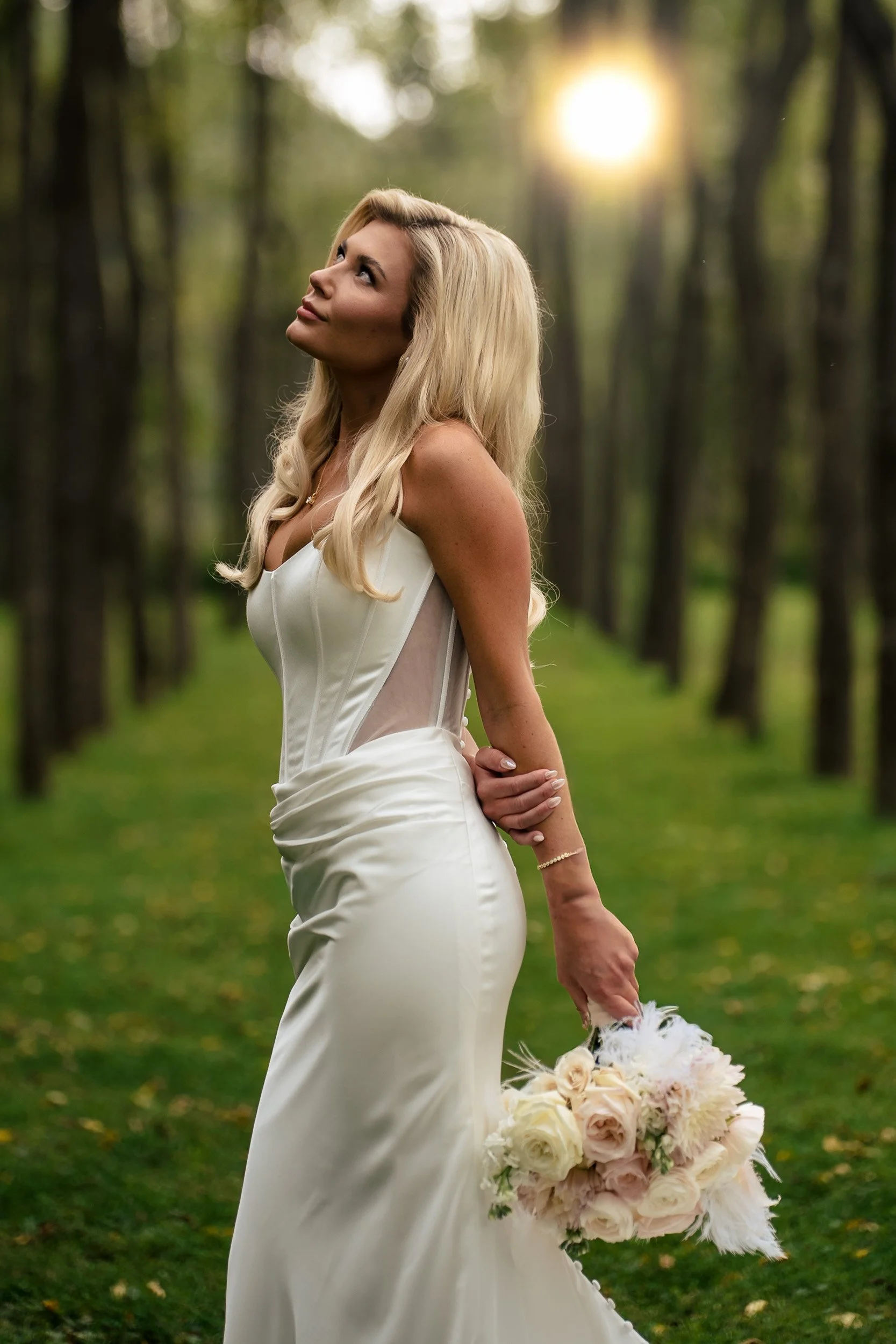Ava holds her bouquet among trees with sunlight behind her.