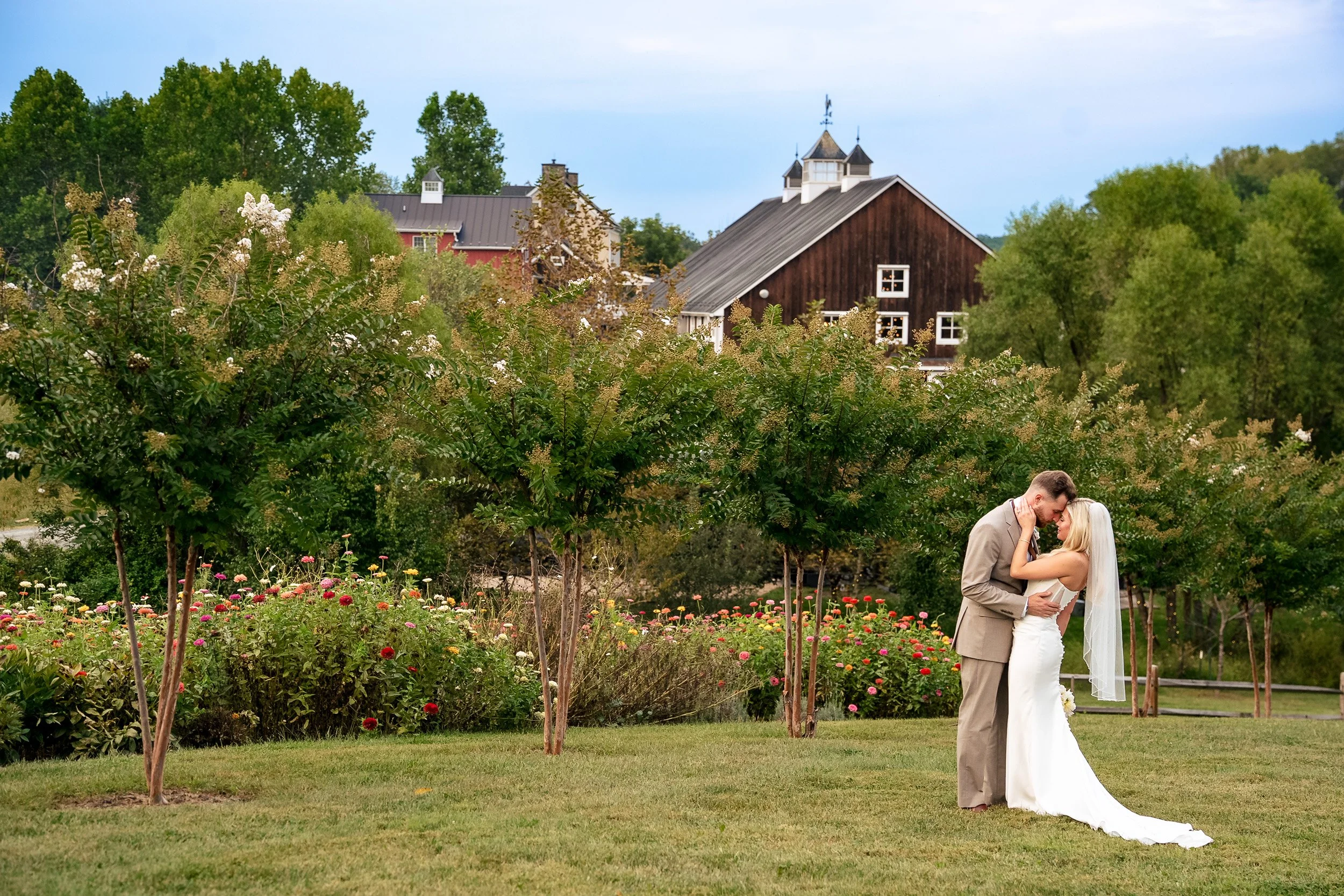 Ava and Jacob kiss on a lawn with flower gardens and a barn-style venue behind them.