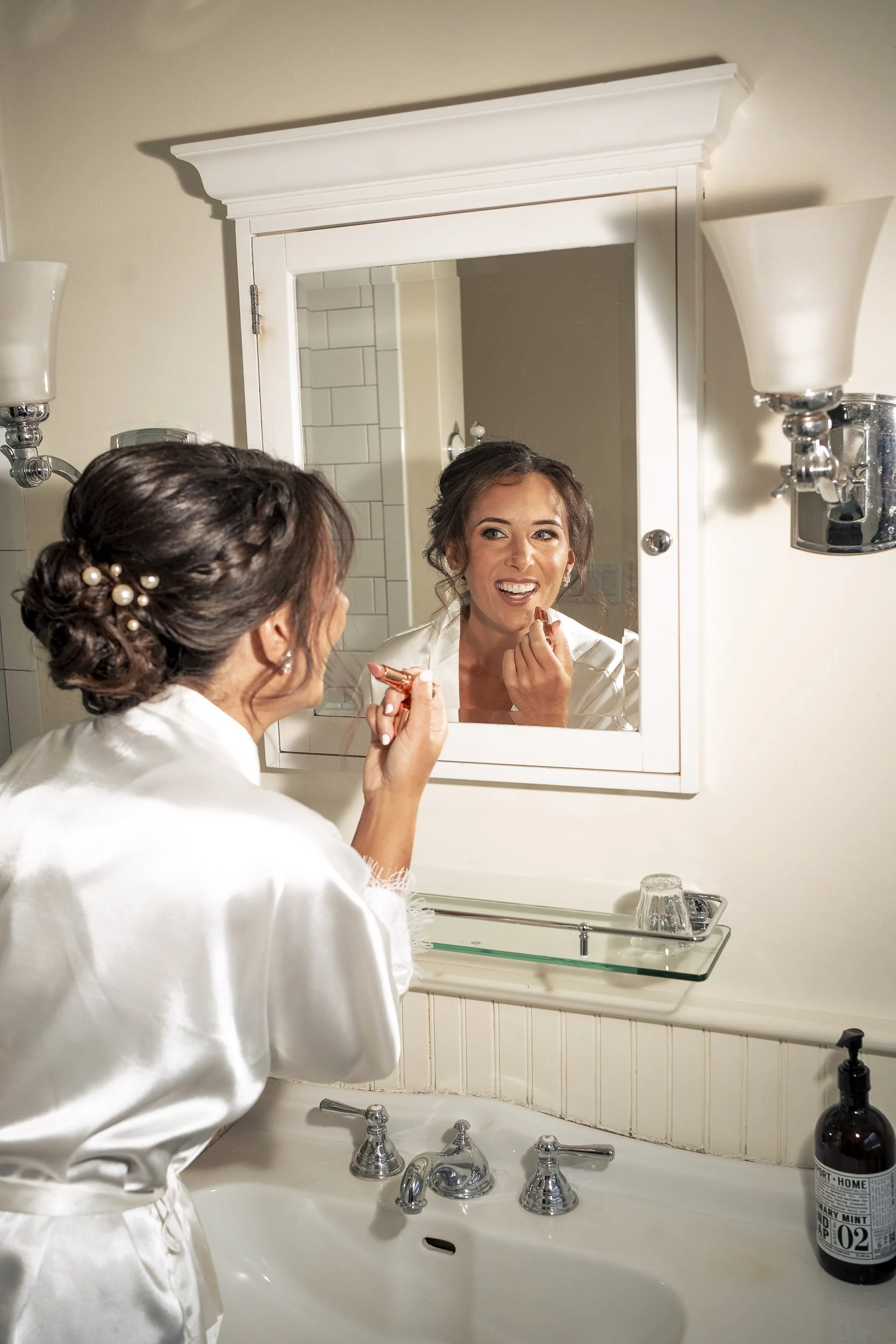 Sarah smiling at her reflection while applying makeup in a white bridal robe.