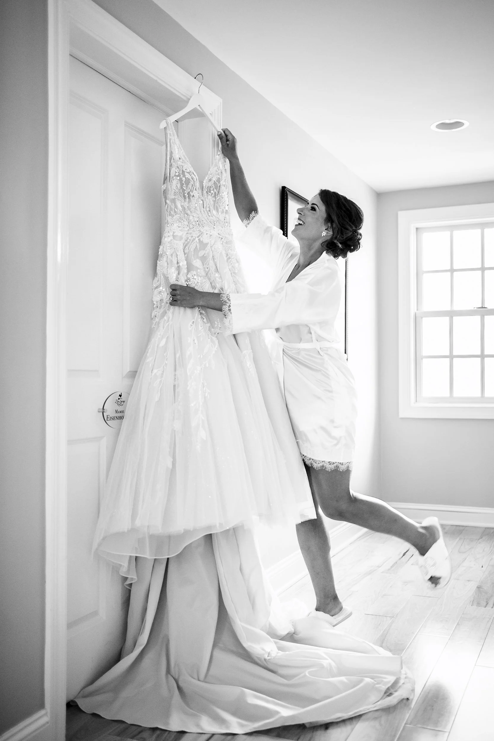 Black and white photograph of Sarah reaching for her hanging wedding dress while lifting one foot behind her.