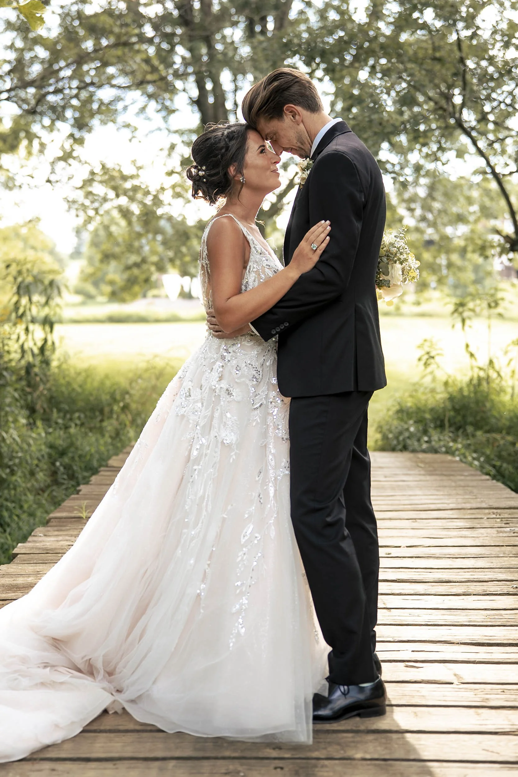 Sarah and Andrew touching foreheads on a wooden bridge beneath leafy trees.