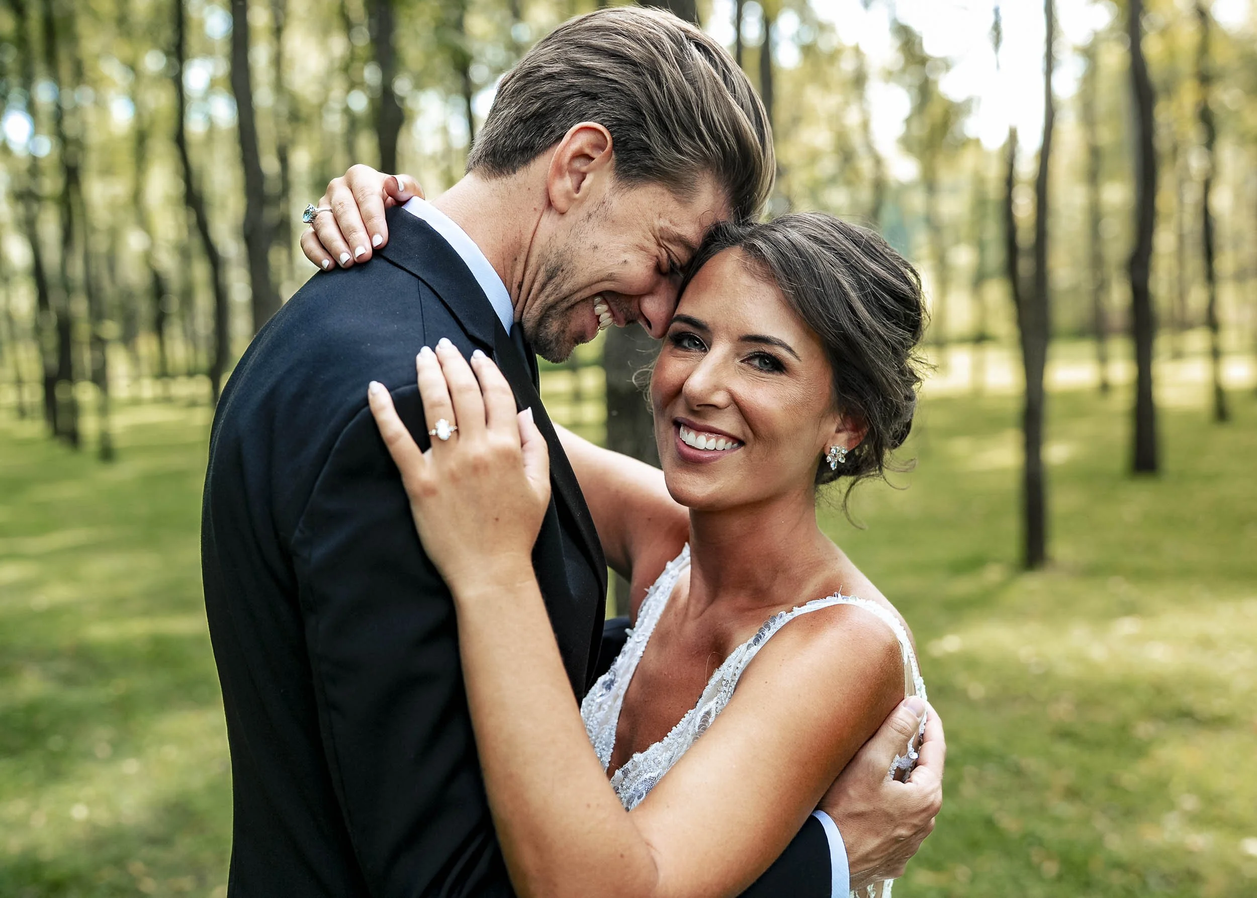 Sarah smiling toward the camera while Andrew rests his forehead against hers during their wooded wedding portraits.