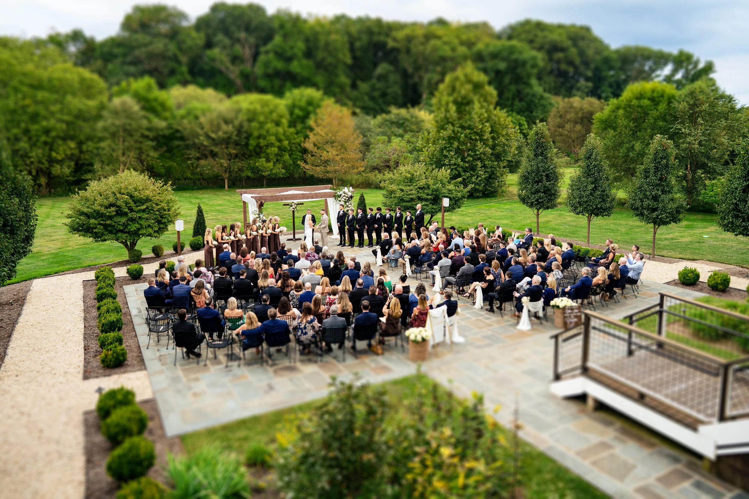 Guests sit around an outdoor ceremony space framed by green hills and trees.
