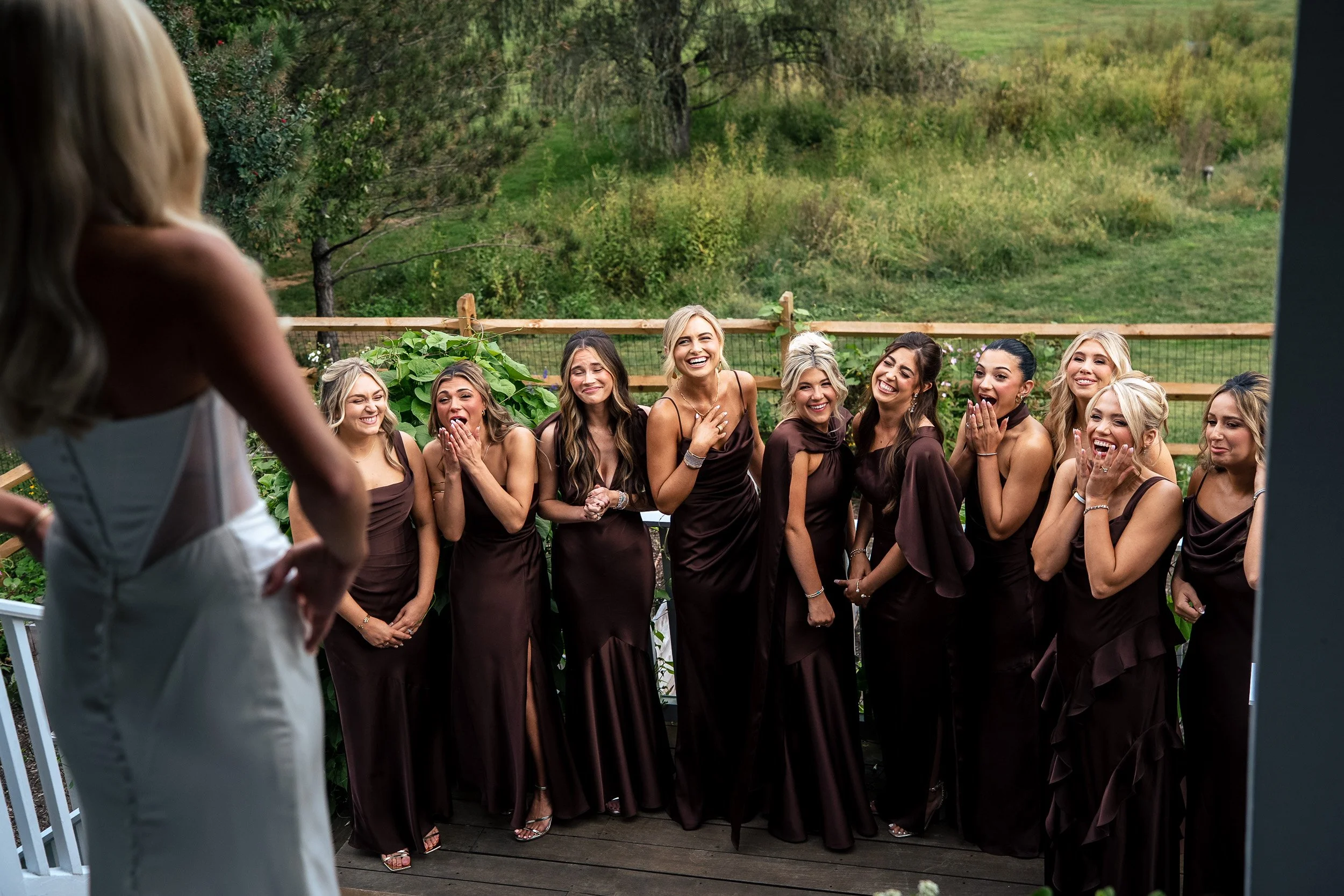 Bridesmaids in chocolate brown dresses smile and react as Ava stands above them on the steps.