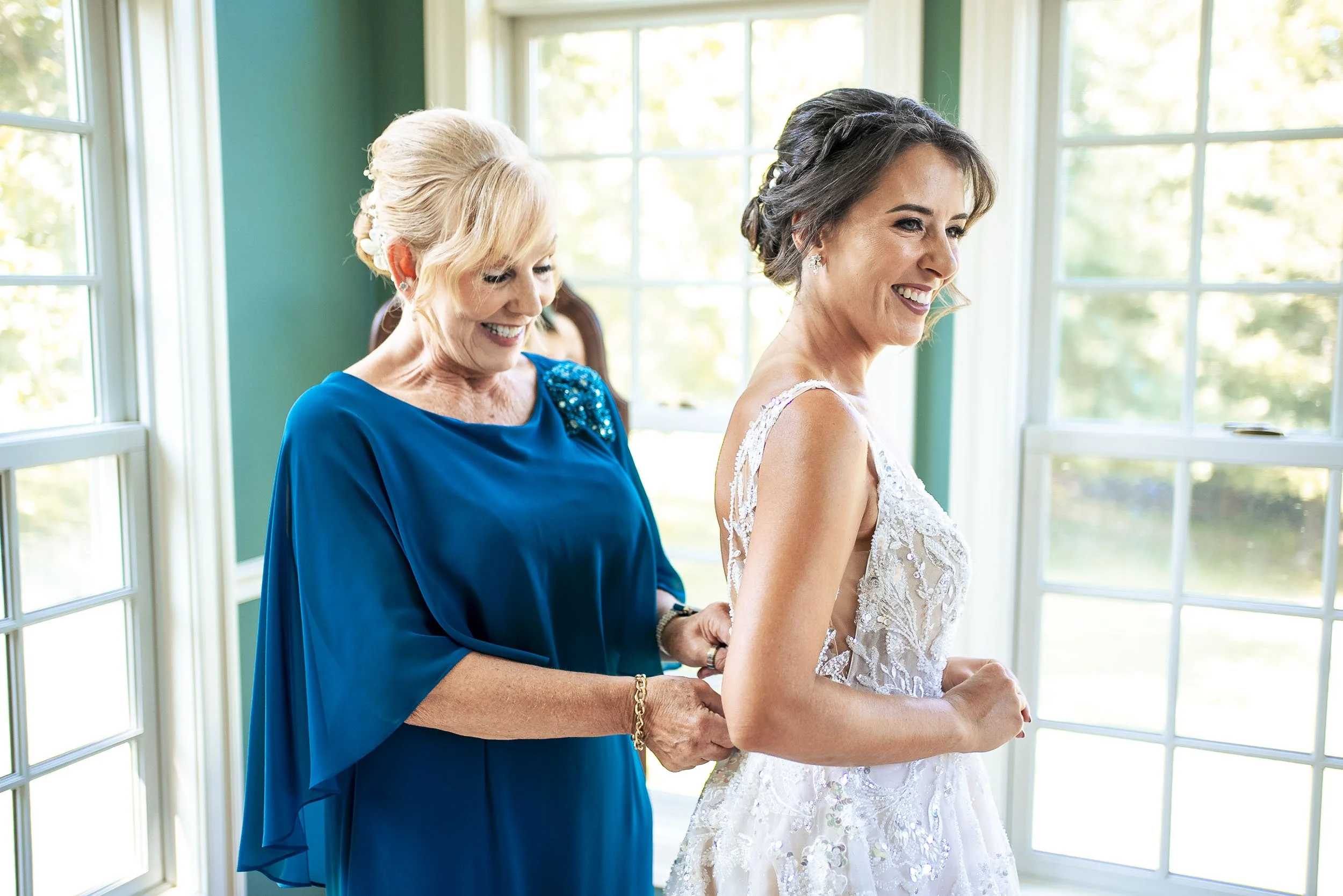 Sarah smiling as her mom in a blue dress helps fasten the back of her wedding gown.