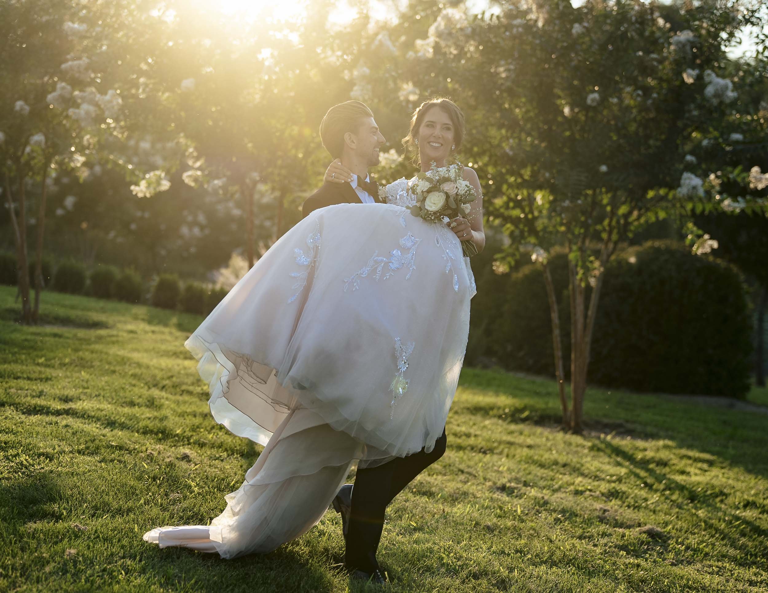 Andrew carrying Sarah in his arms while she smiles and holds her bouquet in golden evening light.